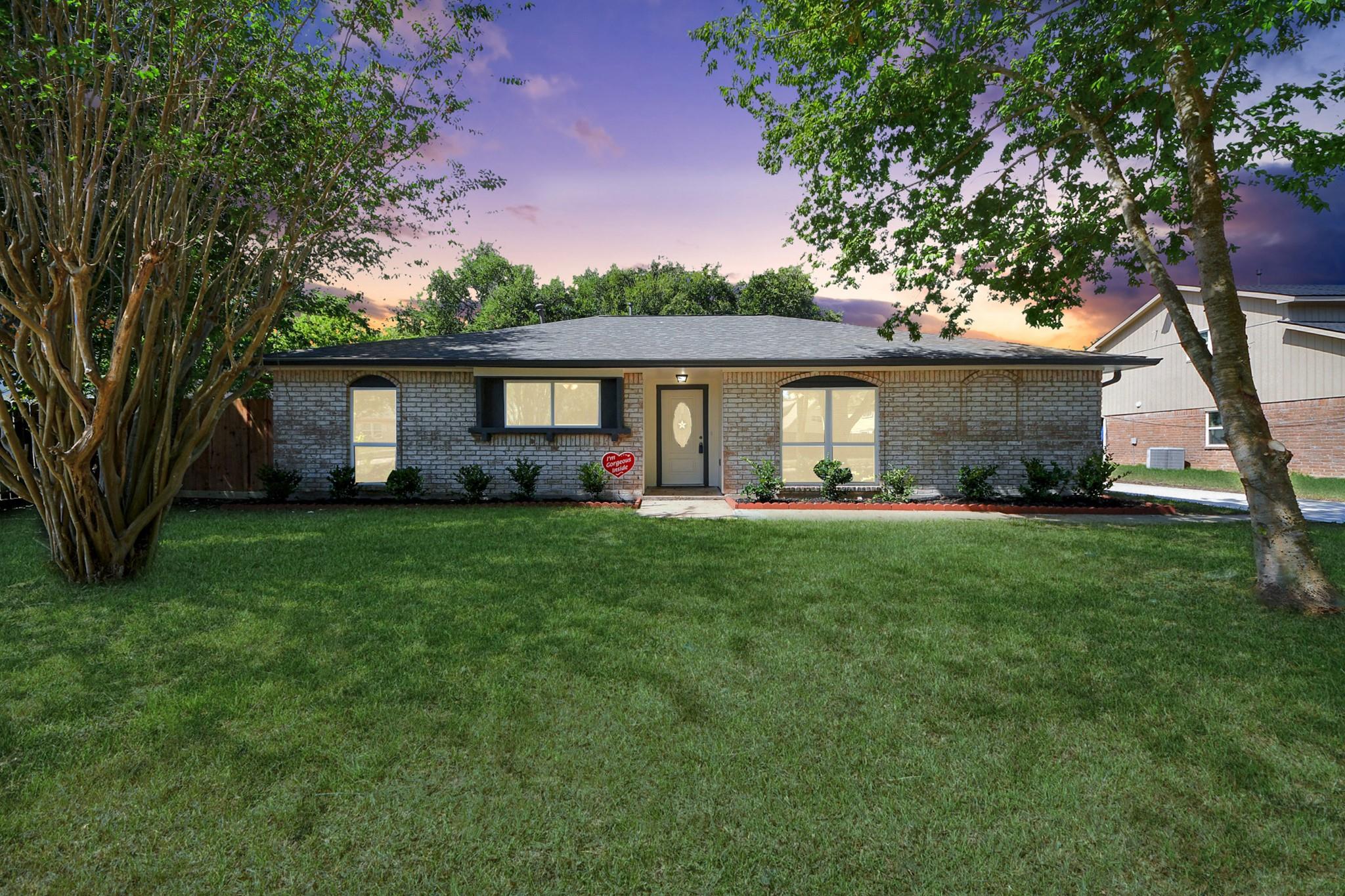 Single-story brick home with a broad lawn, mature trees, and a colorful dusk sky.