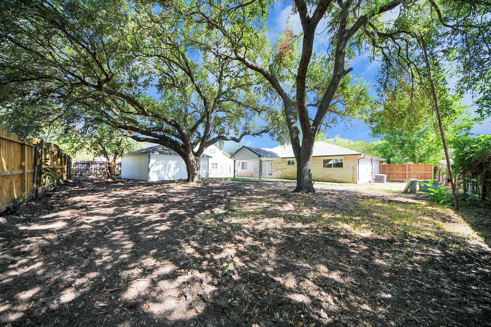 Single-story brick house at the back of a tree-shaded yard, with wooden fencing and a detached white outbuilding.