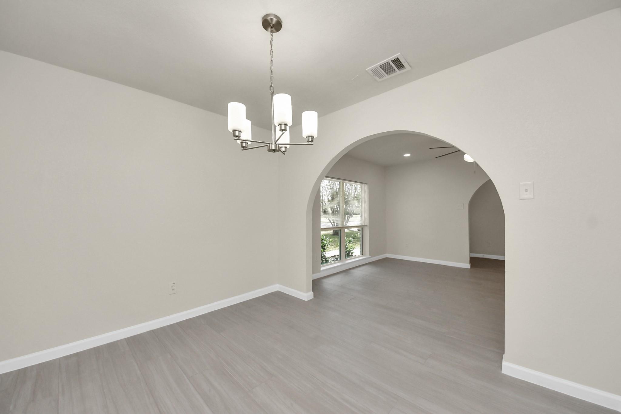 Empty dining room with gray plank floors and chandelier, opening through an archway to a windowed room.