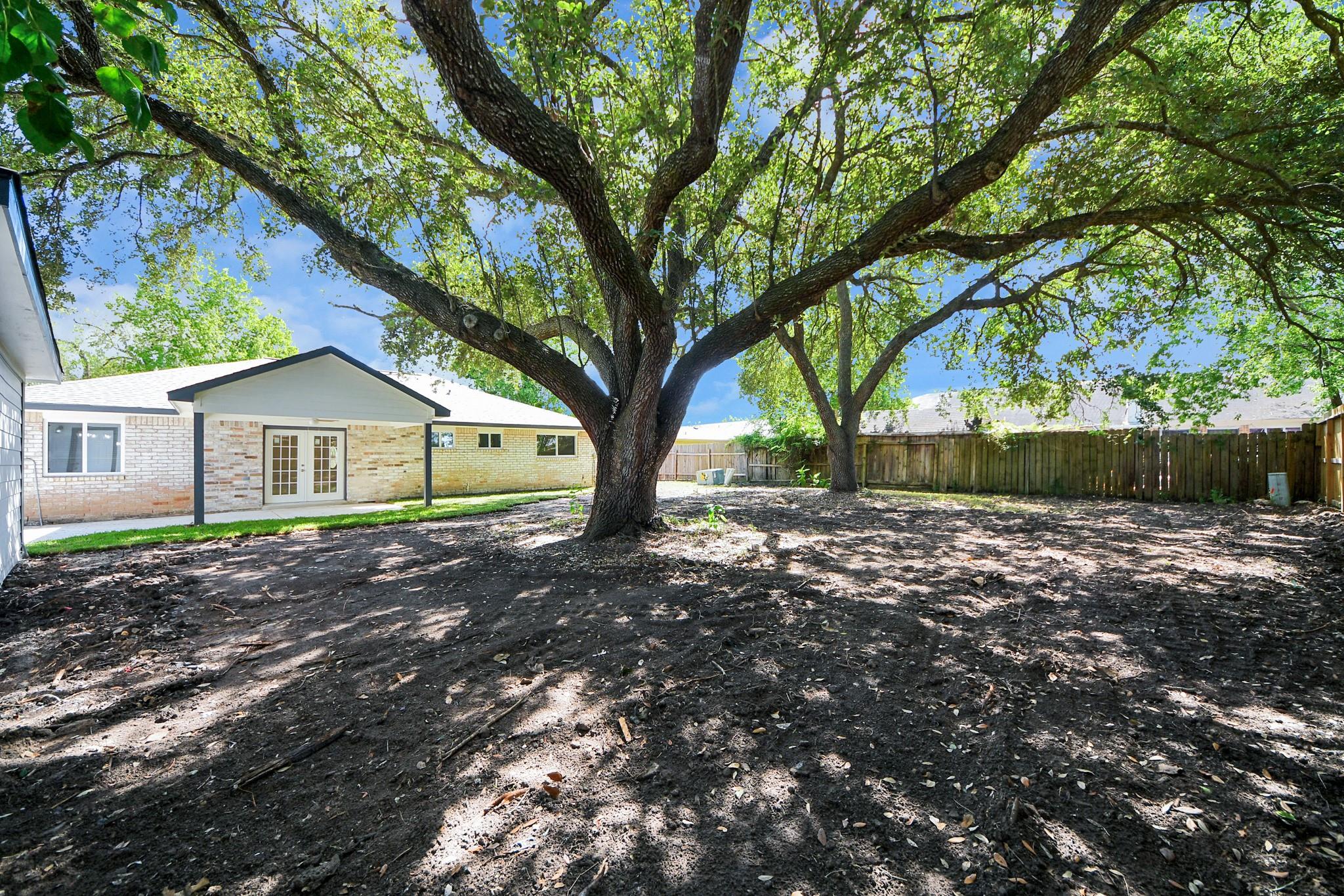 Brick home’s rear patio and doors beneath a covered overhang, beyond a shaded yard with two large trees.