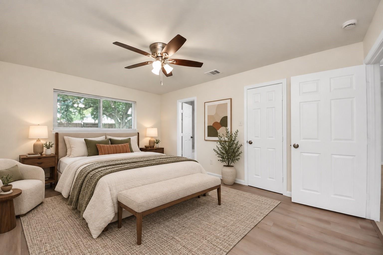 Staged bedroom with neutral bedding, a cushioned bed bench, ceiling fan, and windows with blinds.