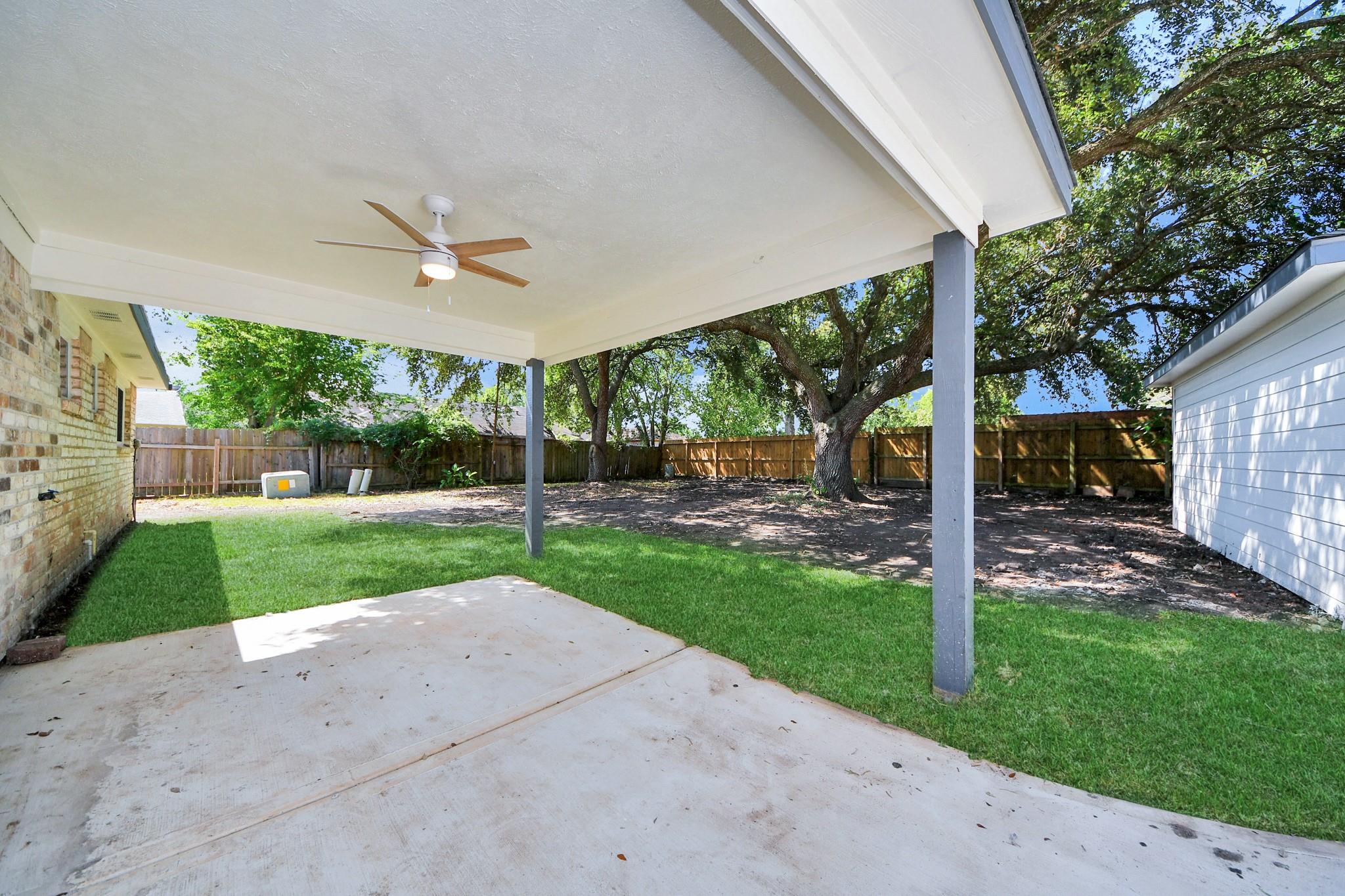 Covered patio with a ceiling fan overlooking a fenced lawn and mature shade trees.