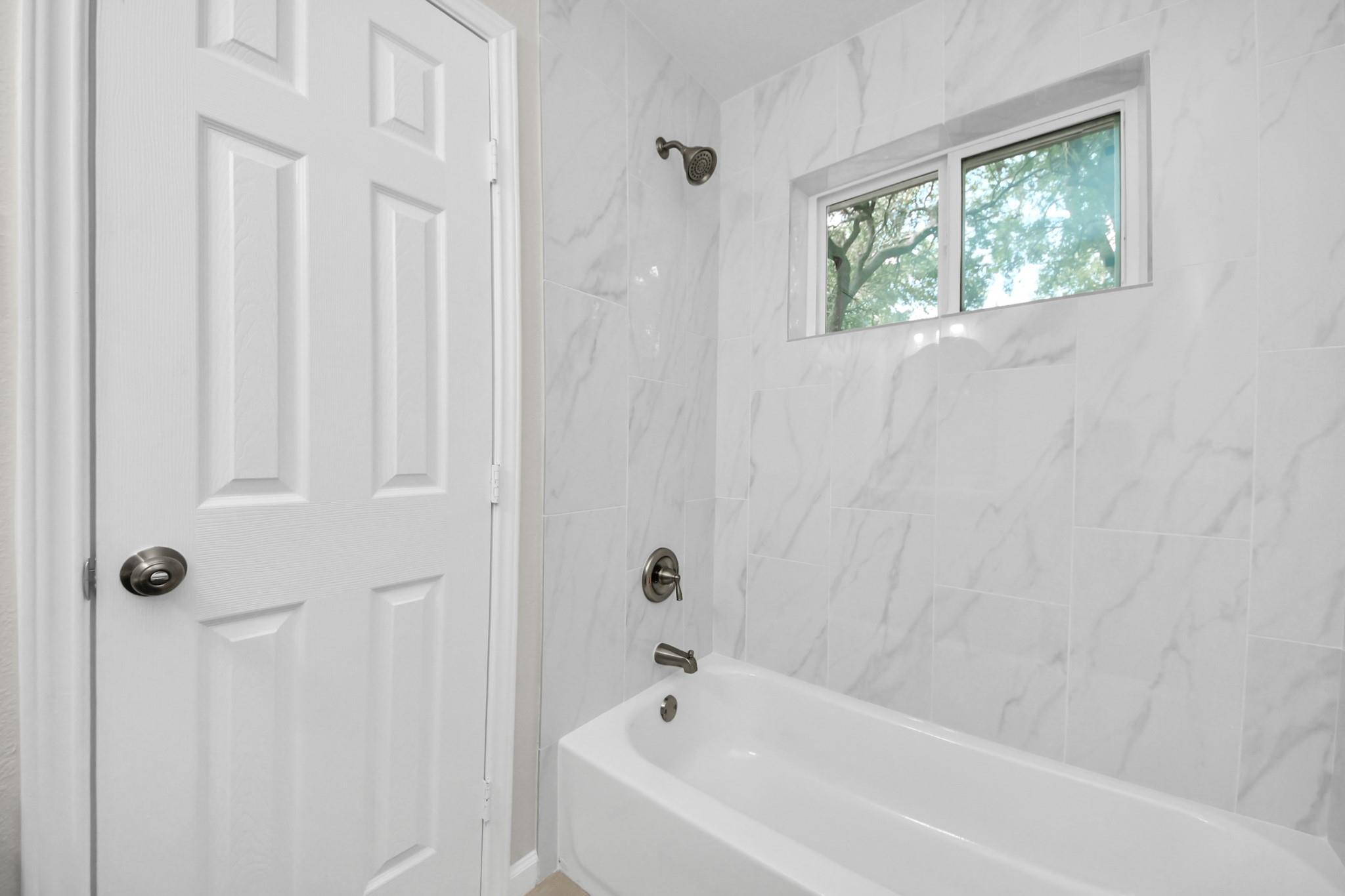 Bathroom with a white tub, marble-look tile, and a small window.