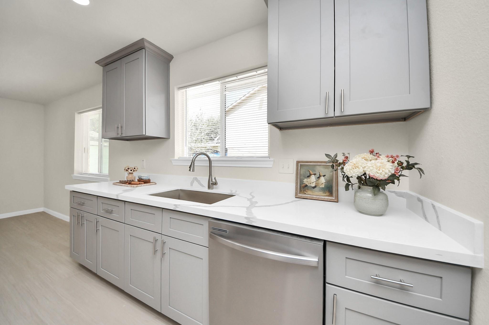 Kitchen with gray cabinets, veined white counters, stainless dishwasher, and sink beneath a window.
