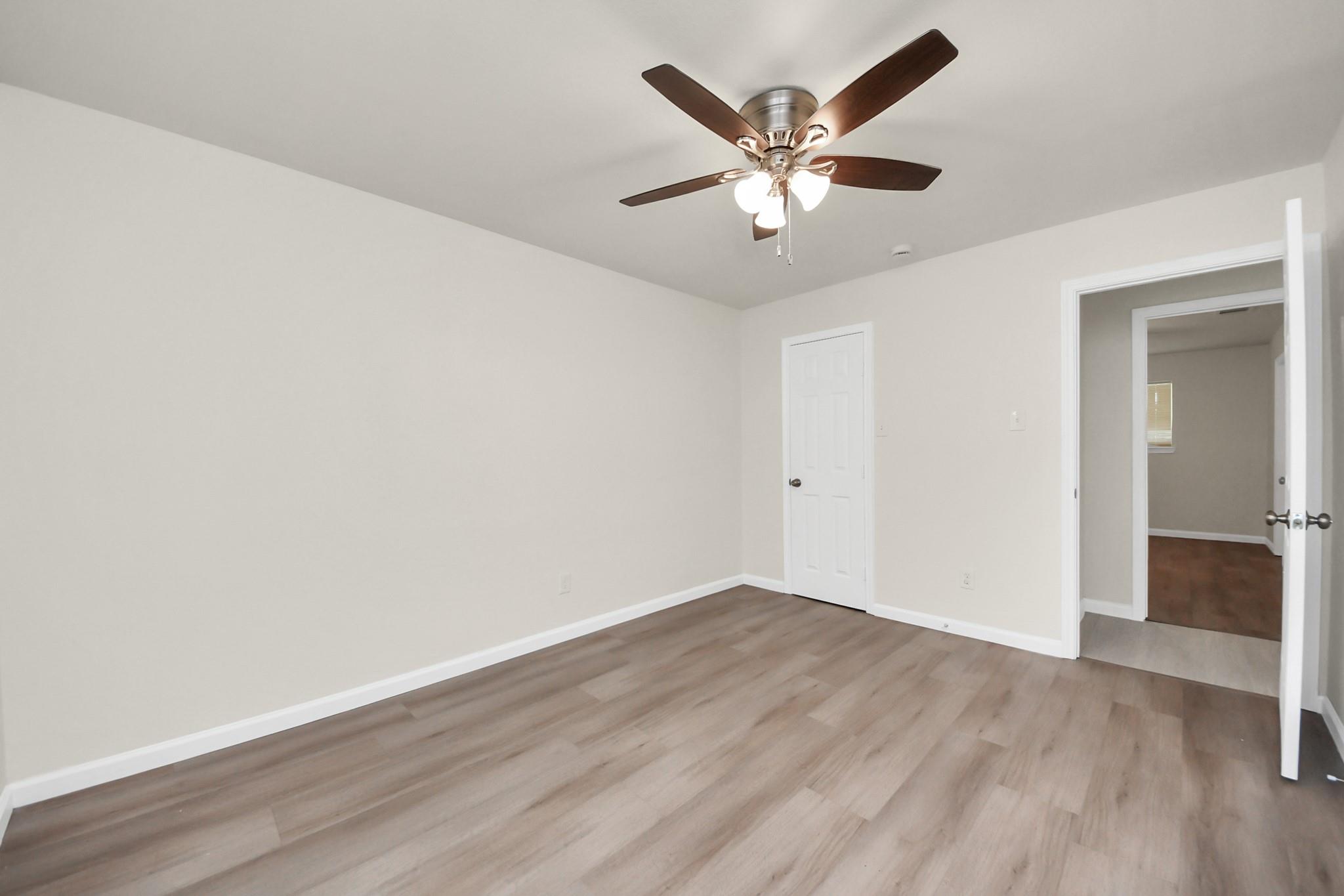 Empty room with gray-brown plank flooring and a lit ceiling fan, viewed toward two doorways.
