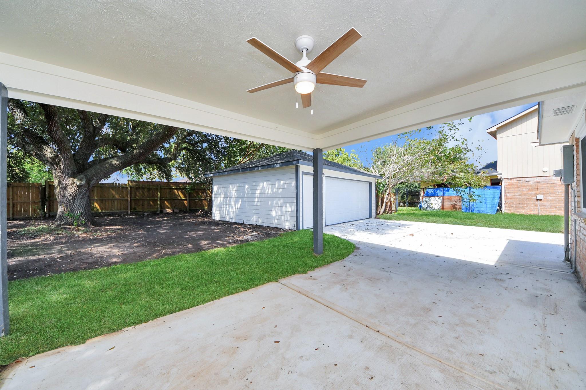 Covered backyard patio with a ceiling fan beside a detached garage and concrete driveway.