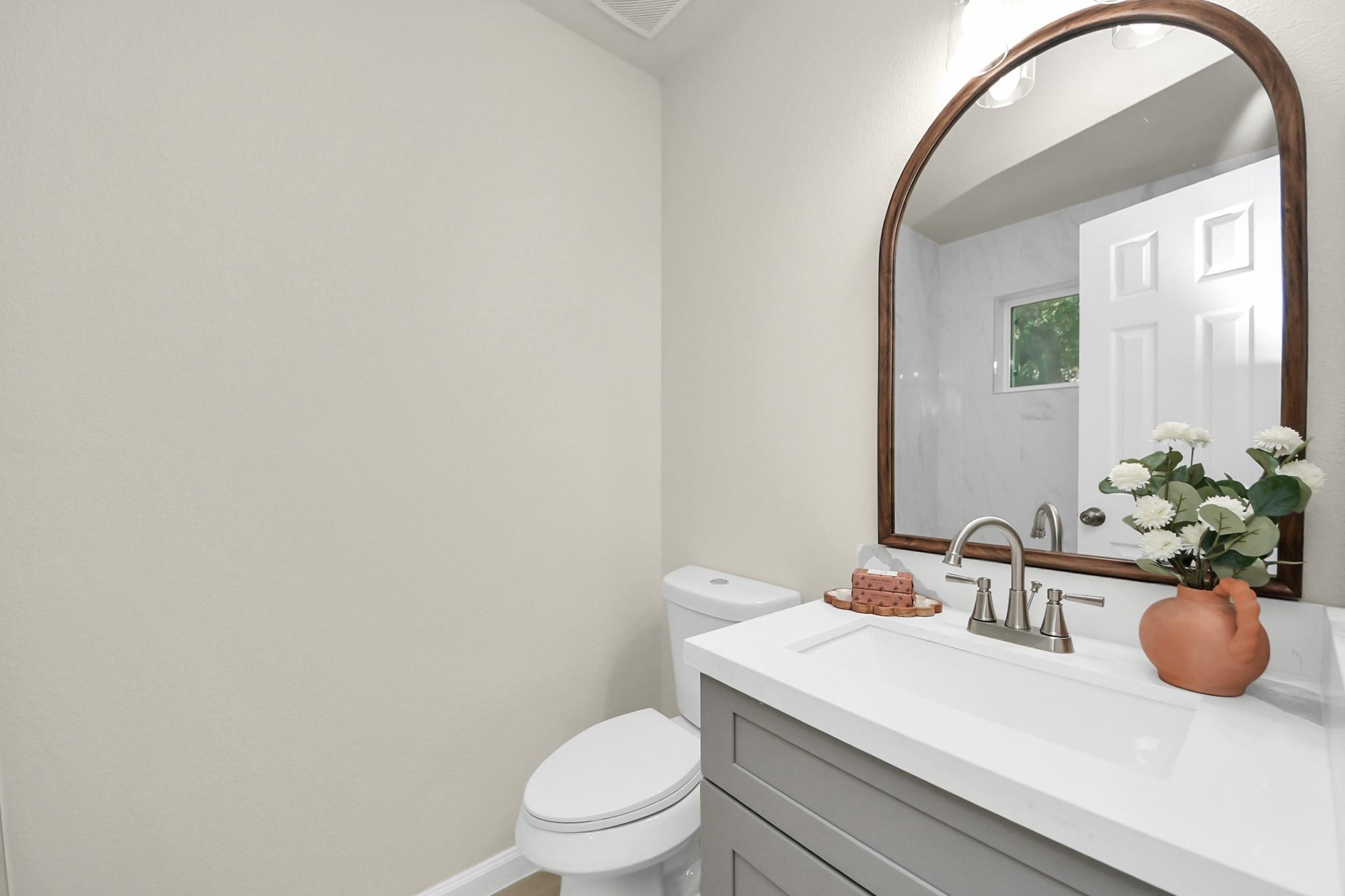 Bathroom with a gray vanity, arched wood-framed mirror, and flowers beside the sink.