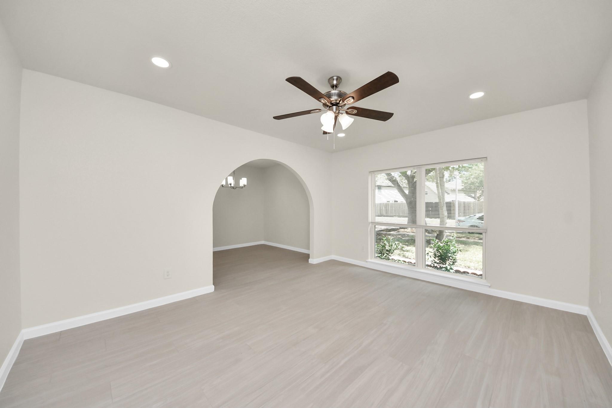 Empty living room with pale flooring, a ceiling fan, a large window, and an arched opening.