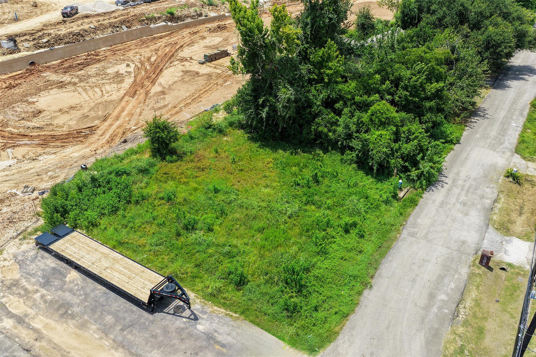 Aerial view of a grassy vacant lot bordered by mature trees and a road, beside a construction site.