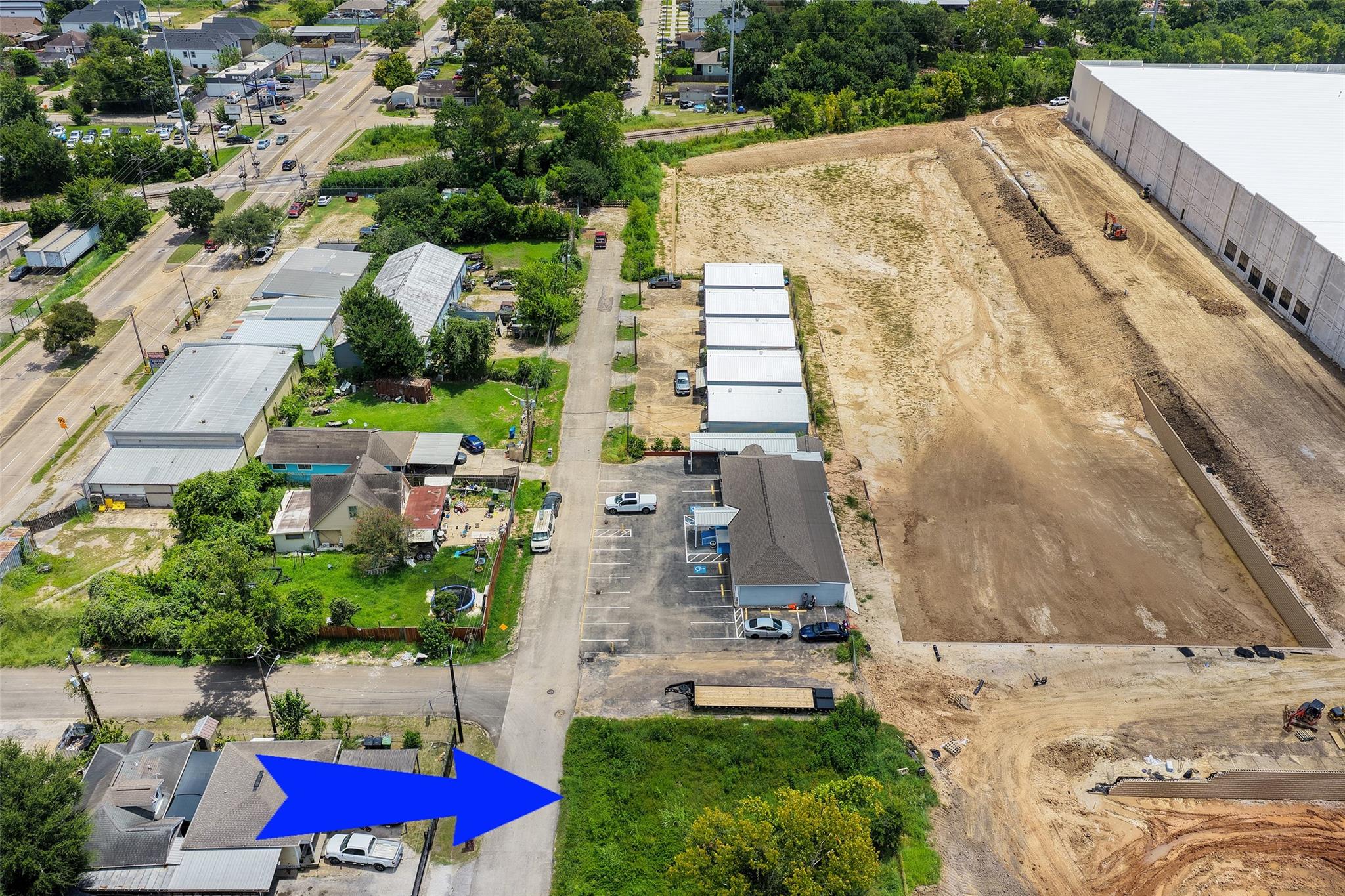Aerial view of a vacant, overgrown lot marked by a blue arrow beside a large construction site.