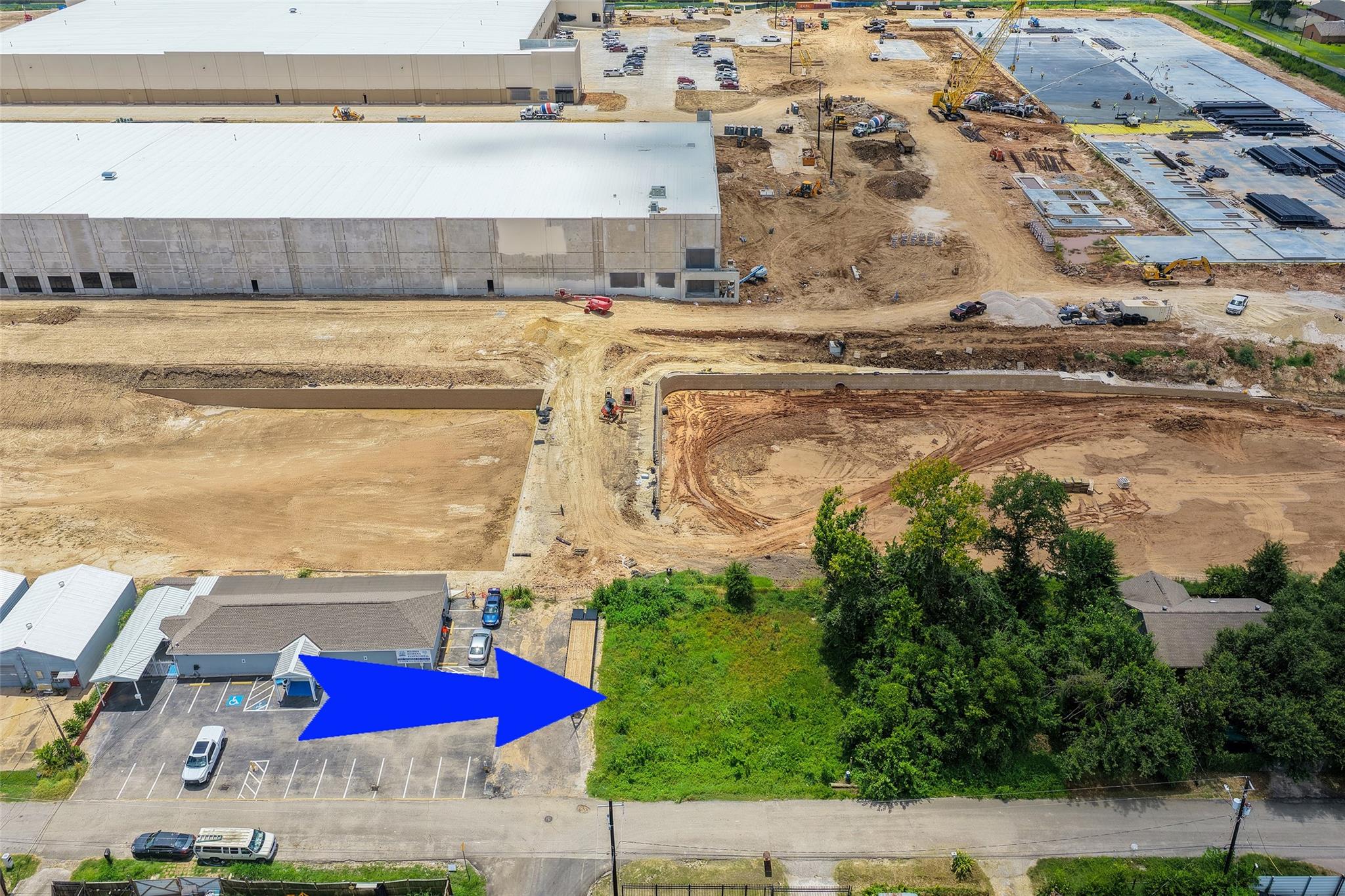 Aerial view of a wooded vacant lot beside a road, marked by a blue arrow next to an industrial construction site.