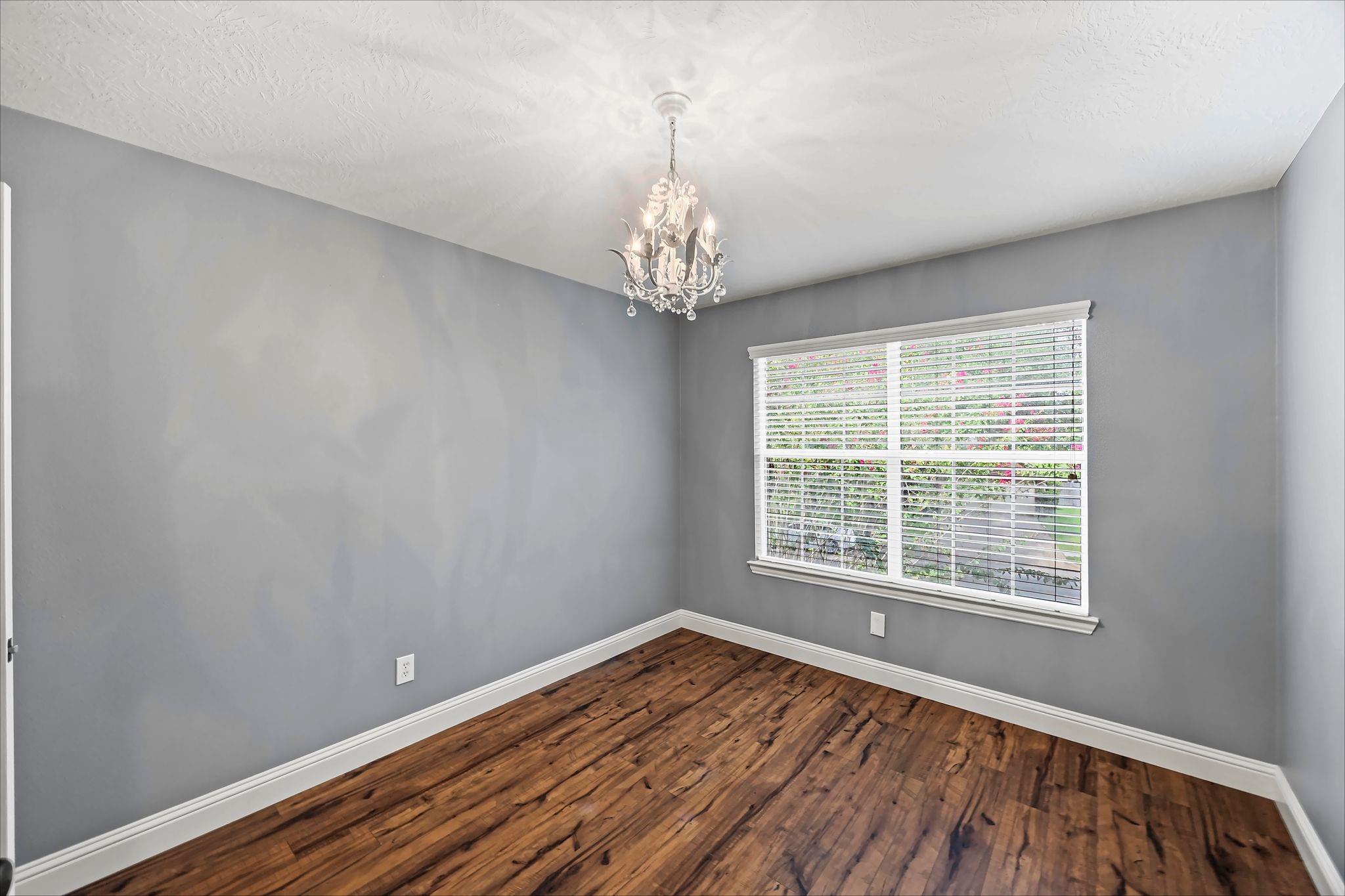Empty gray room with dark wood floors, a crystal chandelier, and a wide window with blinds.