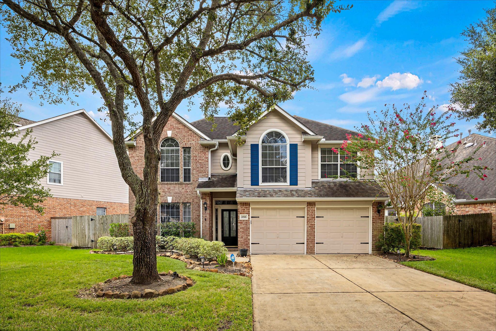 Two-story brick and beige house with blue shutters, double garage, and a large tree in the front yard.