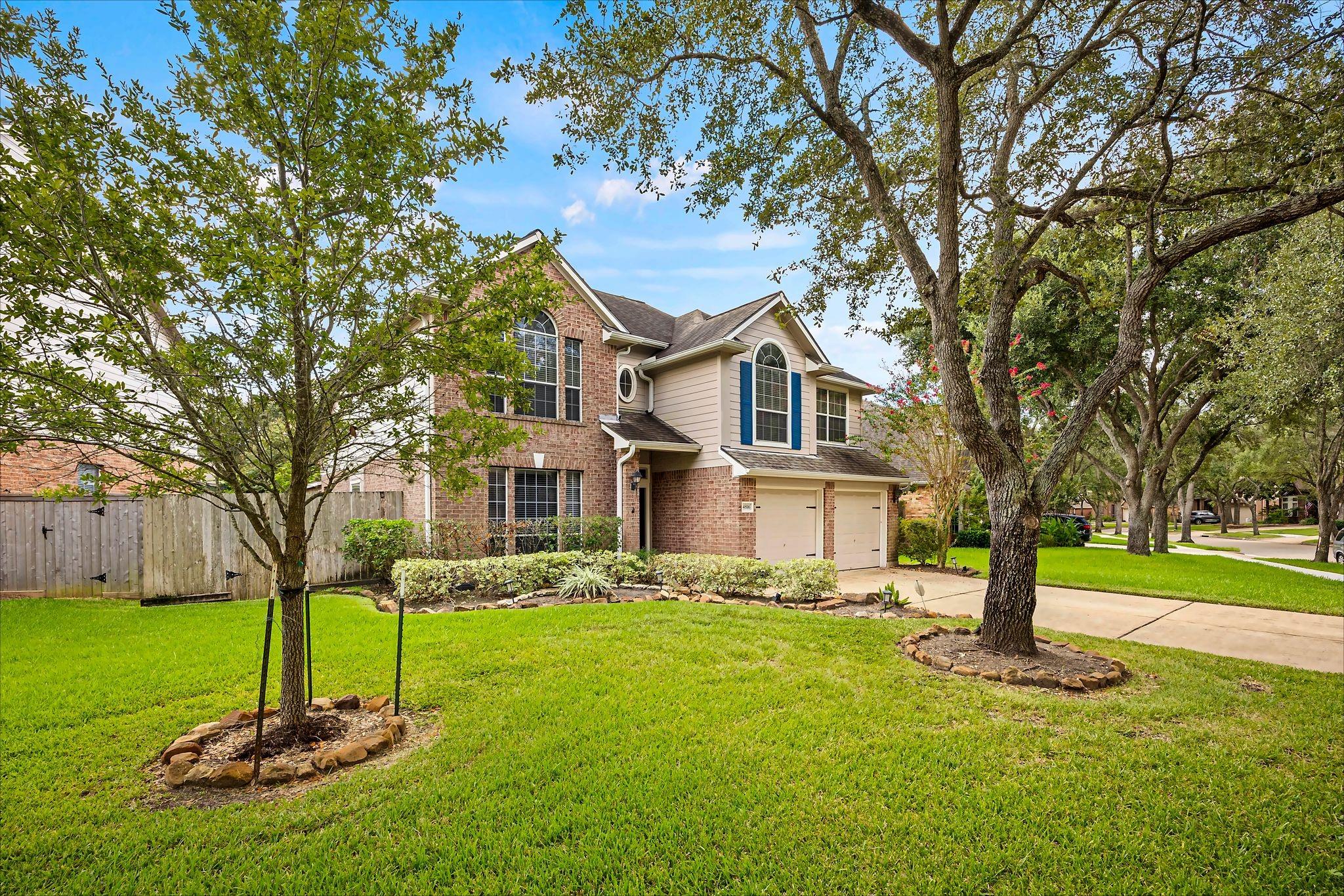 Two-story brick-and-beige home with blue shutters, a two-car garage, mature trees, and a broad green lawn.