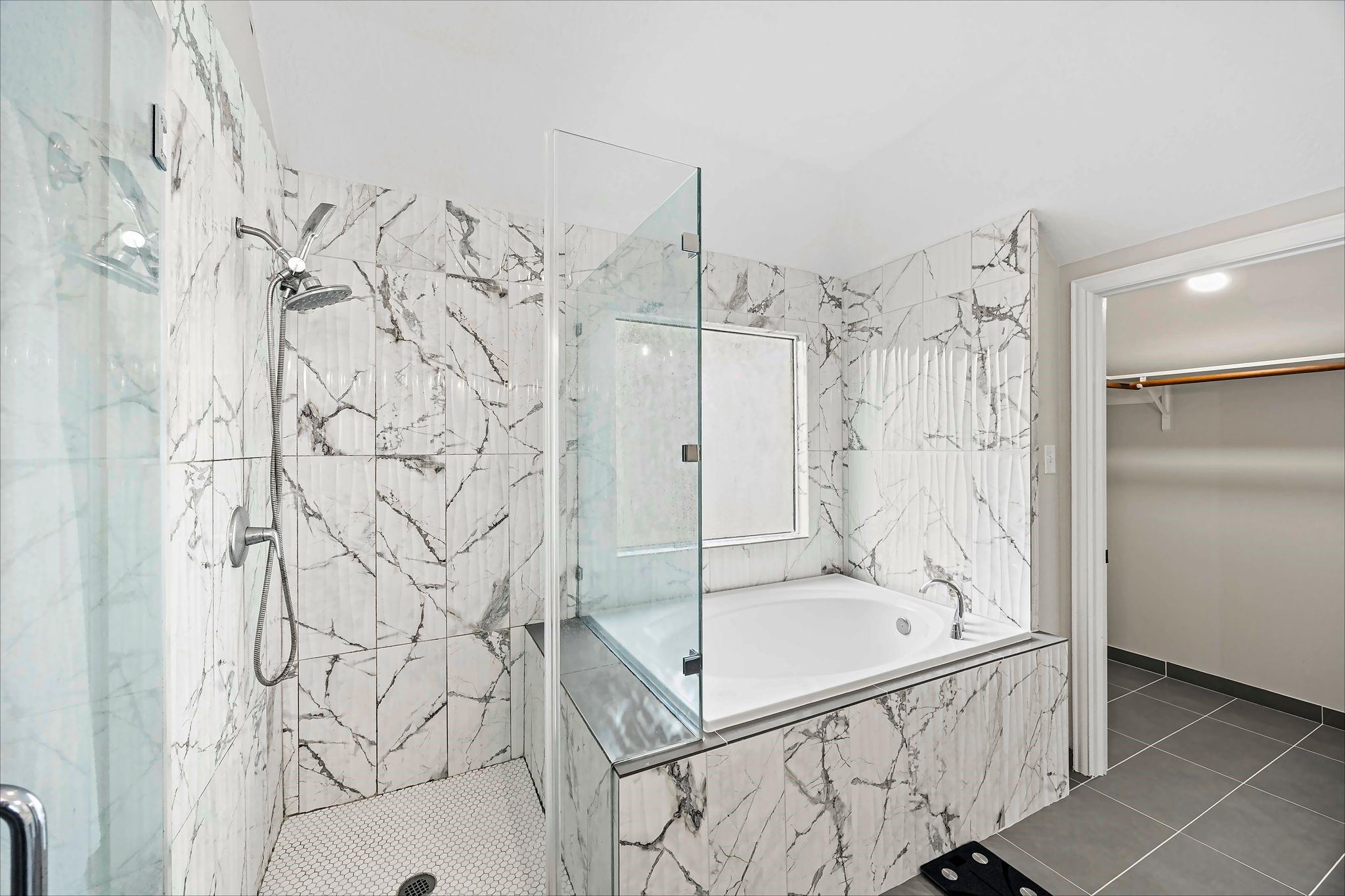 Bathroom with marble-look tiled shower and soaking tub beside an open walk-in closet.