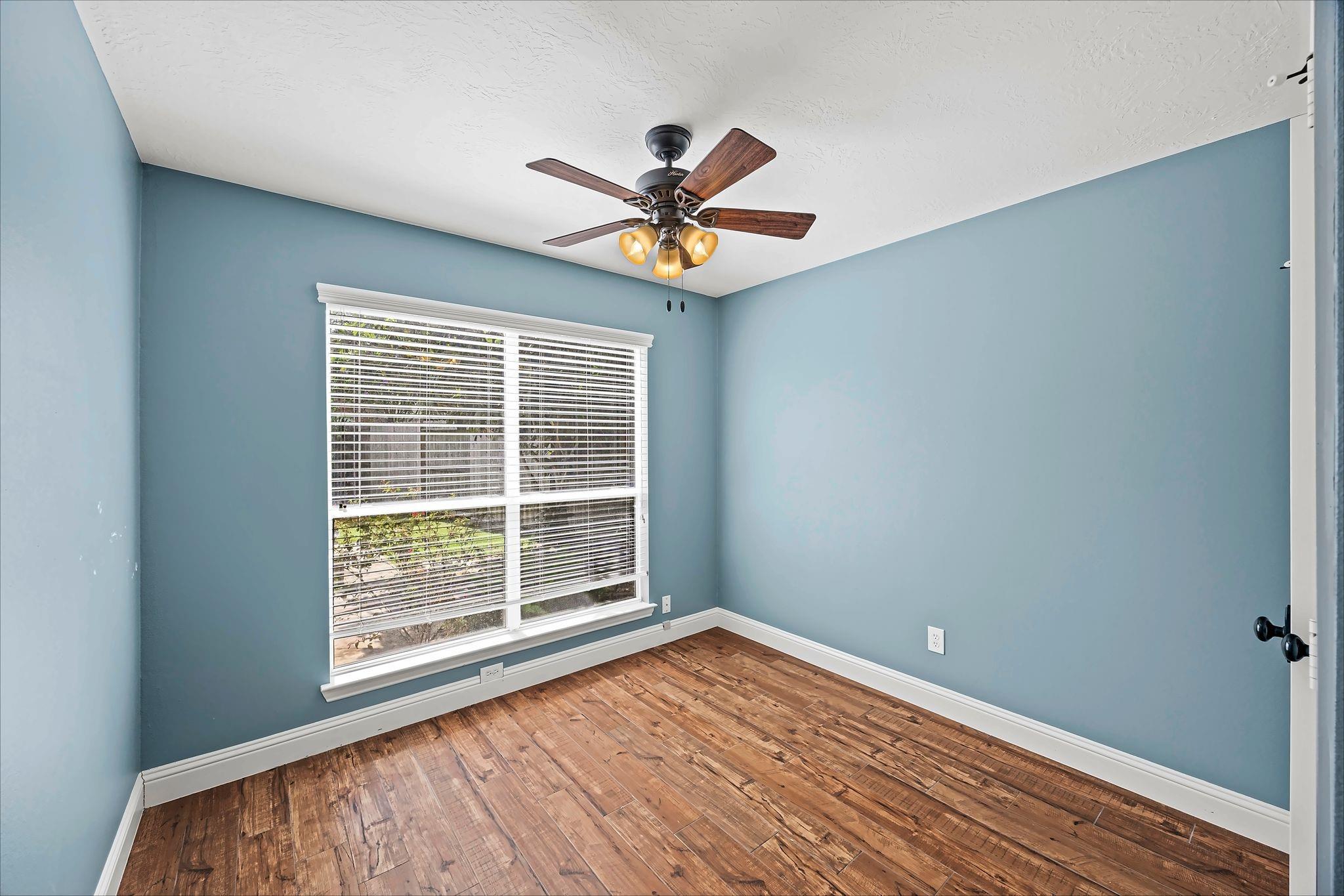 Vacant blue-walled room with wood flooring, a large window, and a ceiling fan.