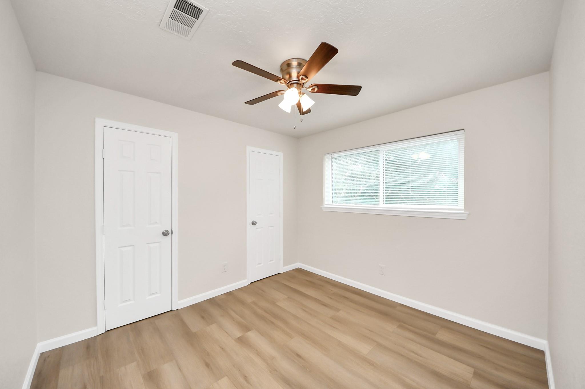 Empty bedroom with wood-look flooring, two white doors, a window with blinds, and a ceiling fan.