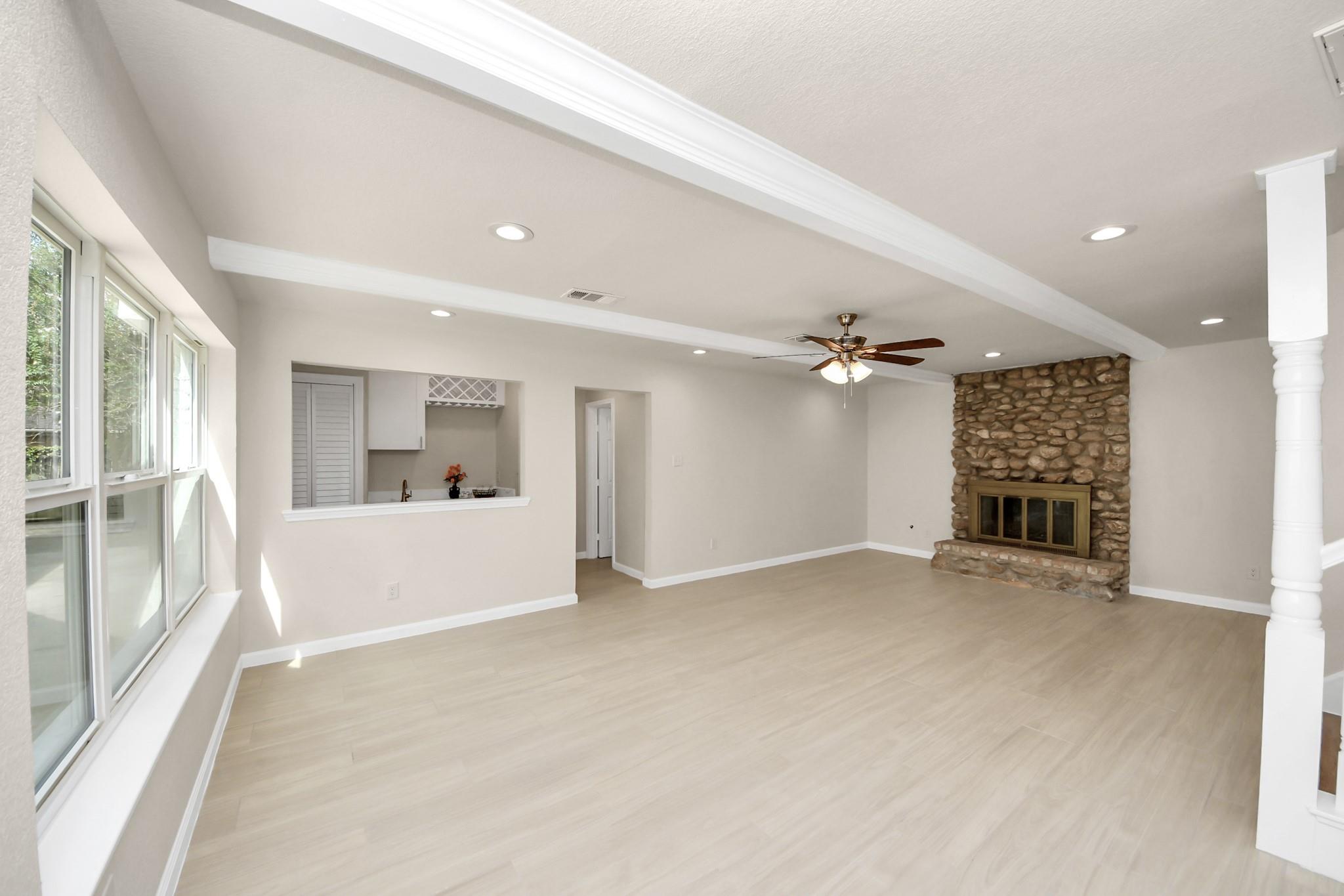 Vacant living room with large windows, pale flooring, and a stone fireplace.