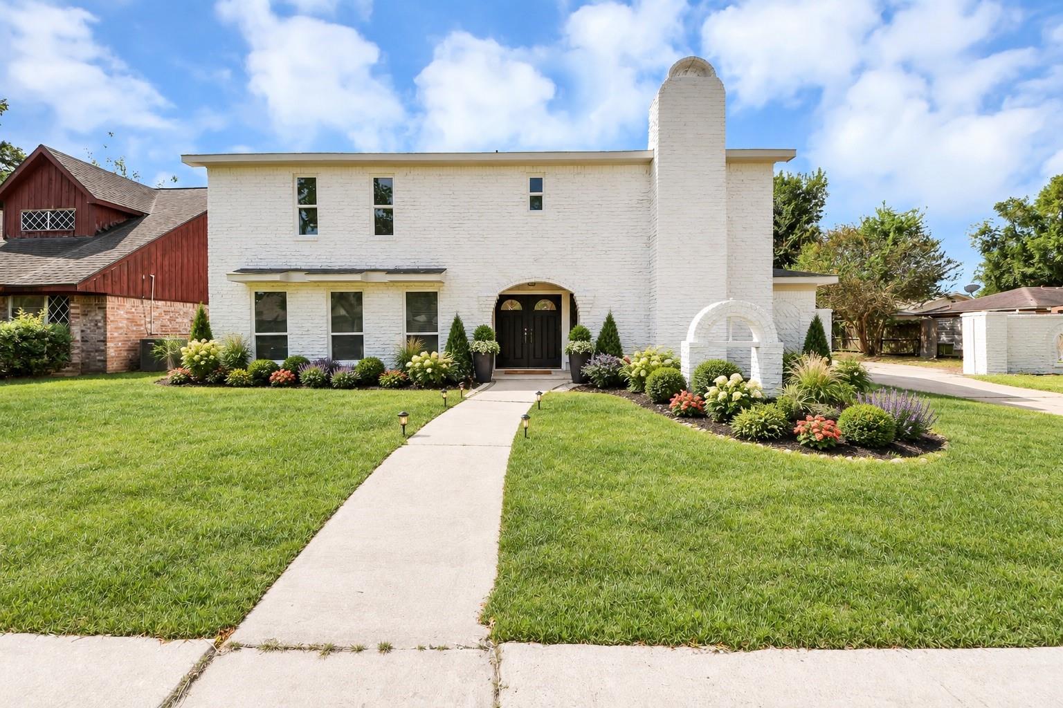 Two-story white brick house with a tall chimney, flower beds, and a walkway through the lawn.