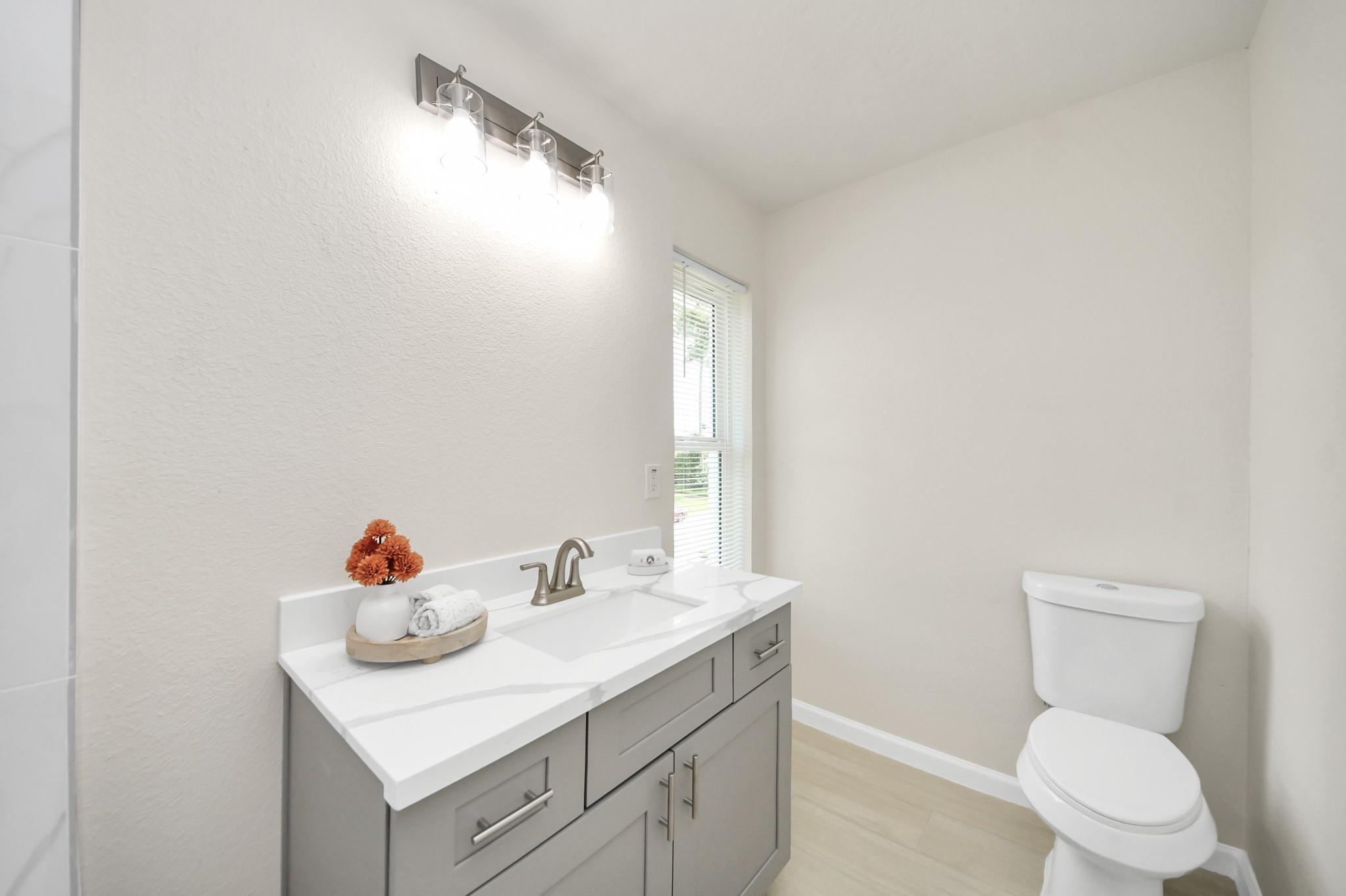 Bathroom with a gray vanity, white countertop, orange flowers, and a toilet beside a narrow window.