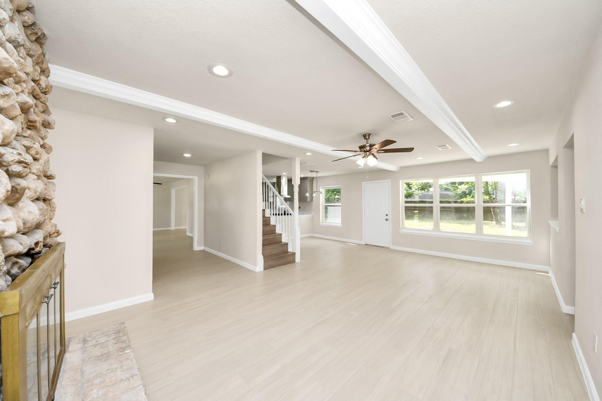 Open-plan living room with a stone fireplace, pale floors, large windows, and stairs beside the kitchen.