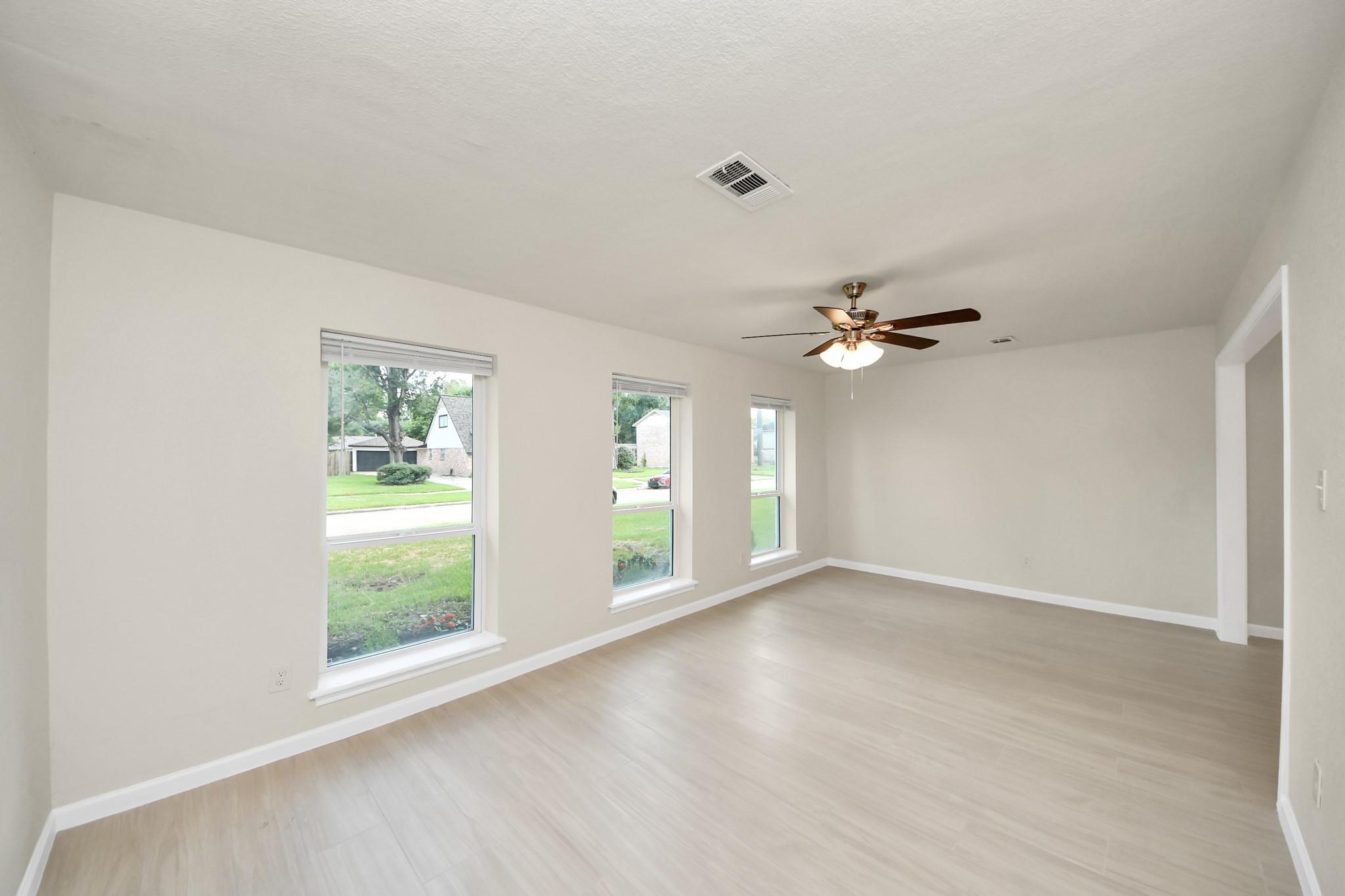 Empty living room with pale wood-look floors, three windows, and a lit ceiling fan.