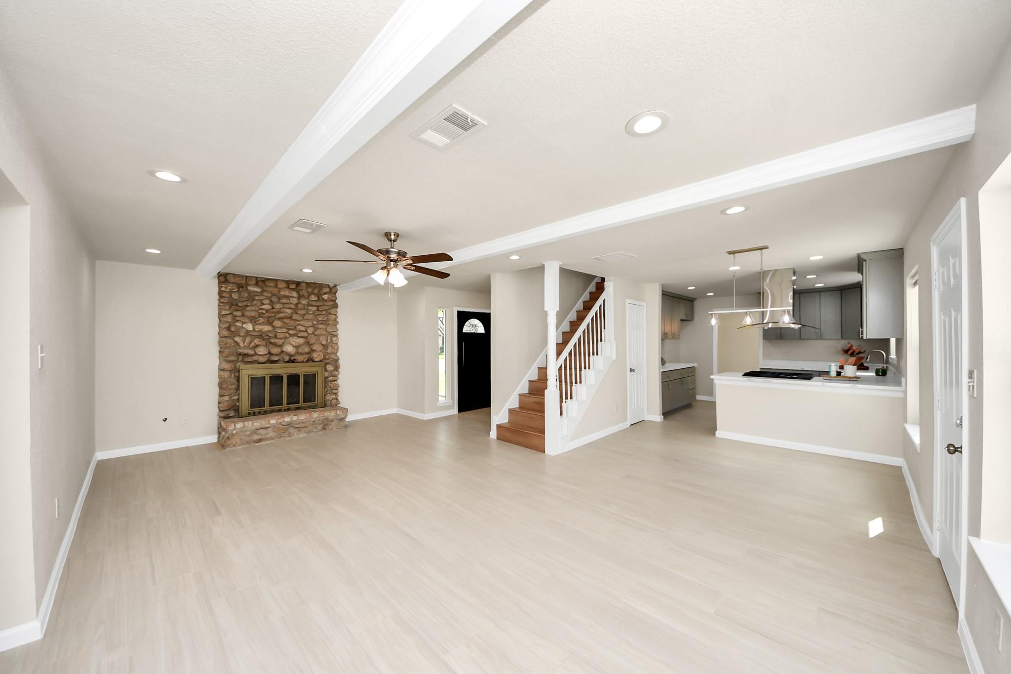 Open-plan living room with a stone fireplace, staircase, and kitchen with gray cabinets.