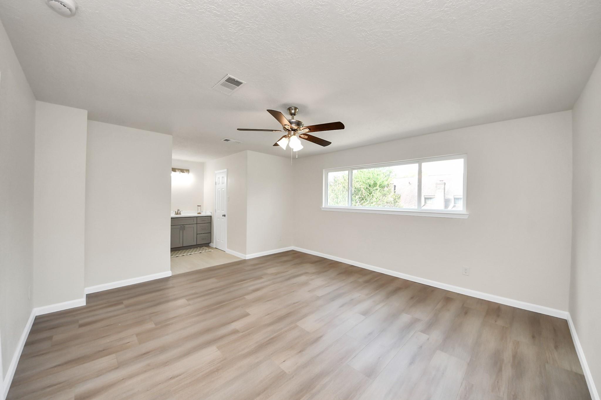 Empty bedroom with wood-look flooring, a ceiling fan, and a wide window beside an open vanity area.
