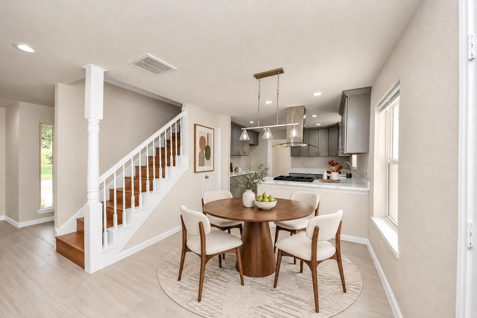 Dining area with a round wood table beside a gray-and-white kitchen and wood staircase.