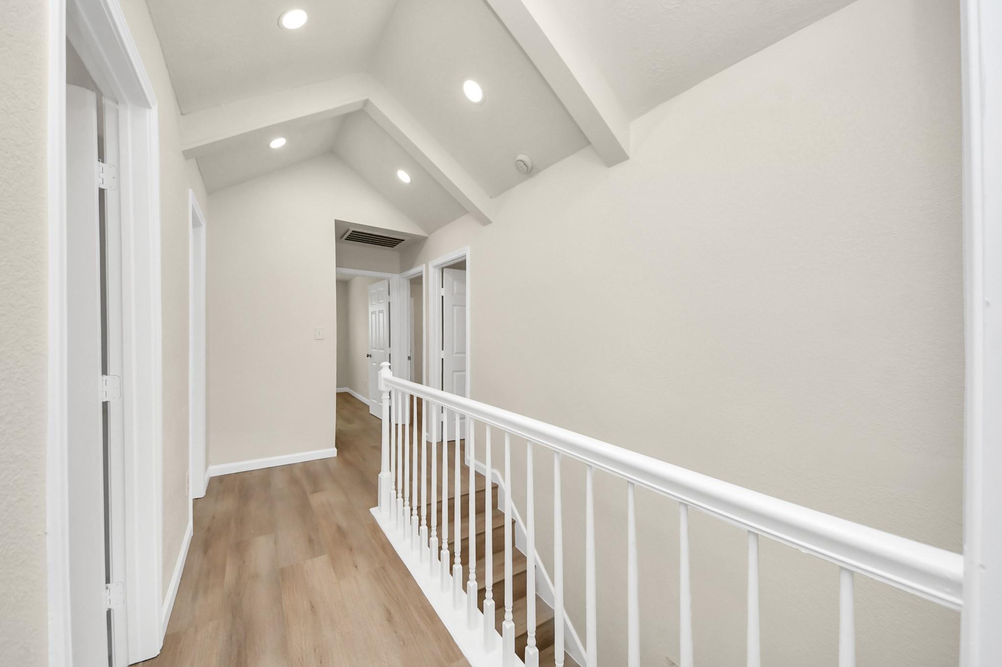 Upstairs hallway with a white stair railing, light wood floors, and a vaulted beamed ceiling.