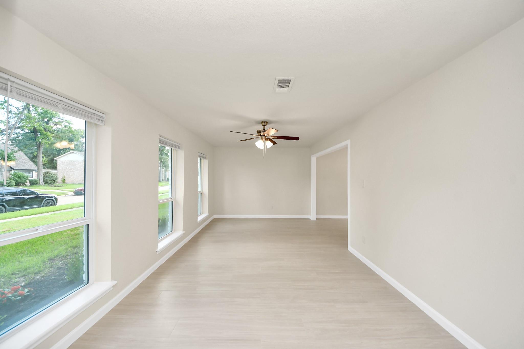Vacant room with light wood-look floors, a ceiling fan, and tall windows overlooking the lawn.