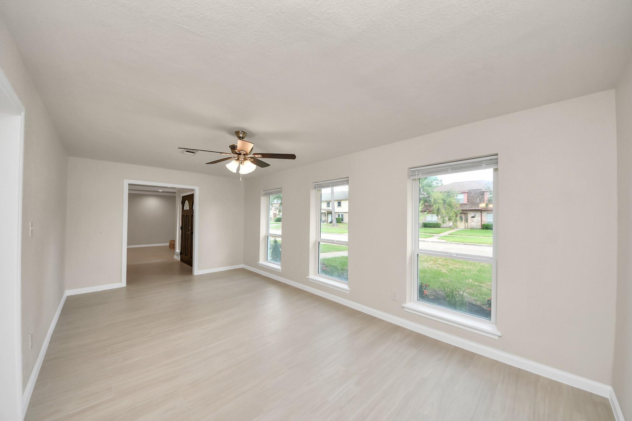 Empty living room with light flooring, a ceiling fan, and three front-facing windows.