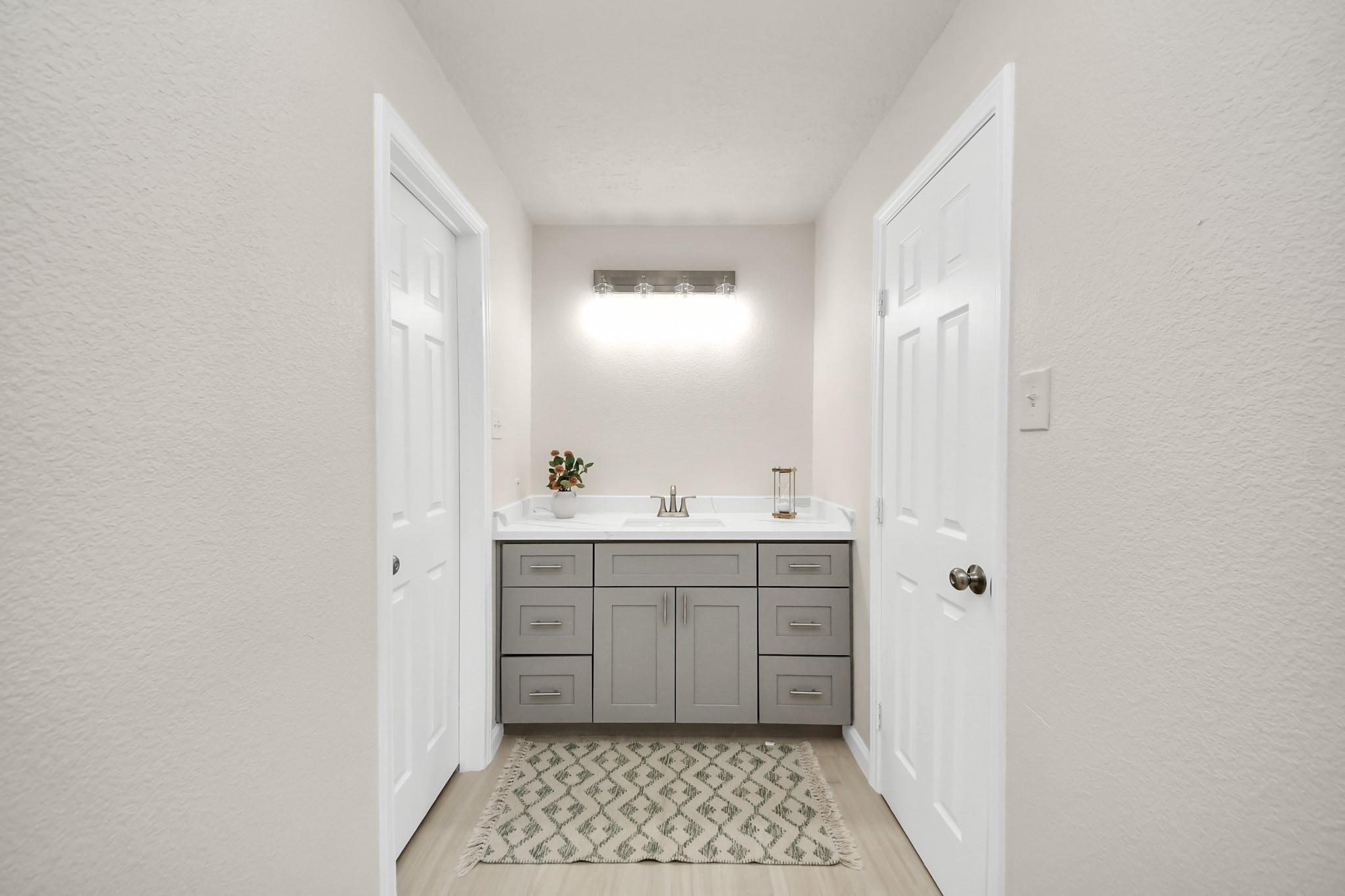 Bathroom vanity with gray cabinets, white countertop, wall light, and patterned rug.