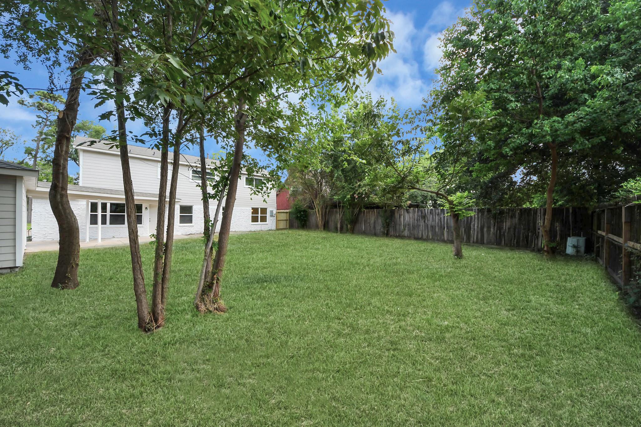 Grassy backyard with mature trees, a wooden privacy fence, and the rear of a white two-story house.