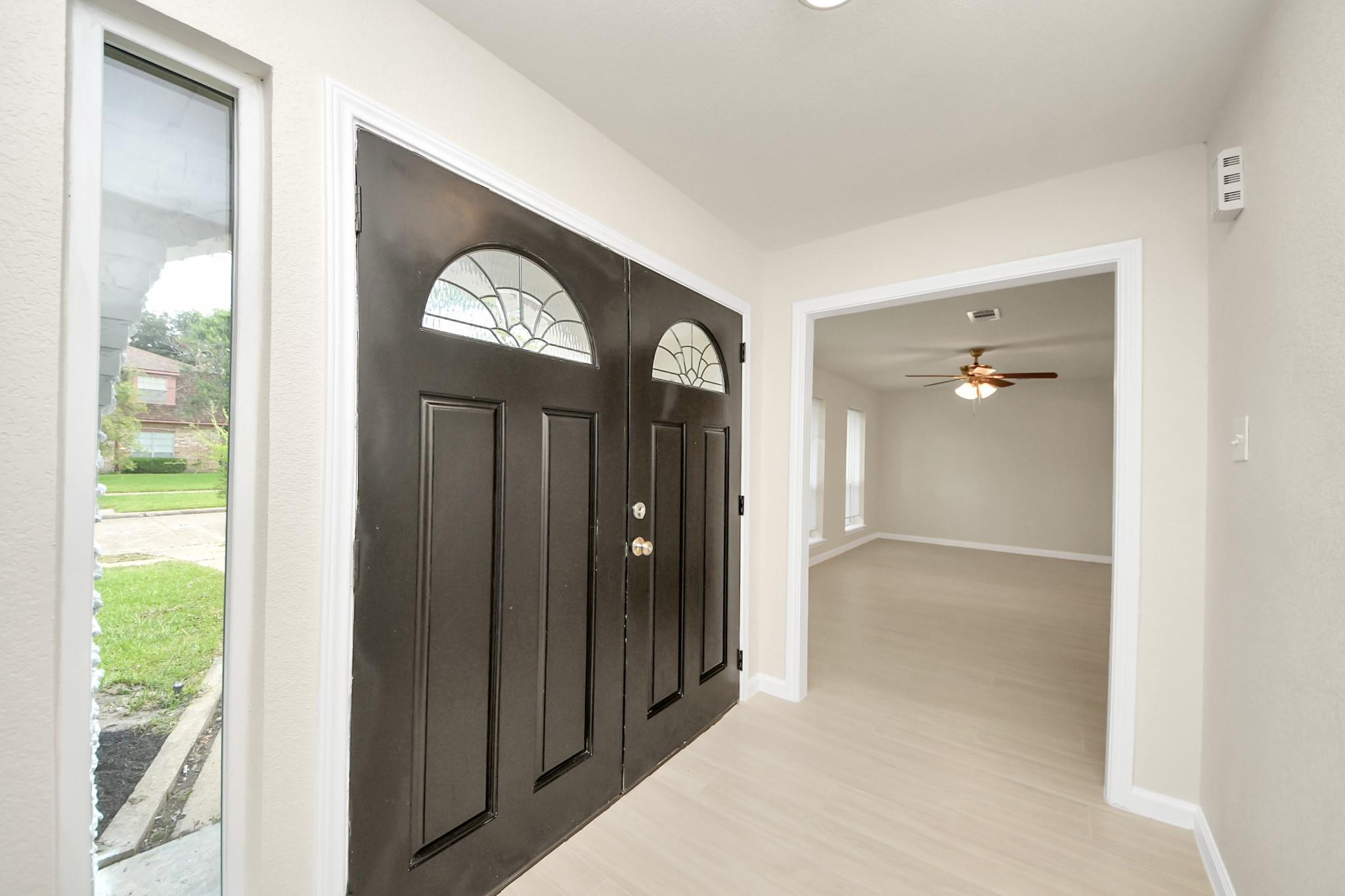 Entry foyer with dark double doors and decorative glass, opening to a vacant room with a ceiling fan.