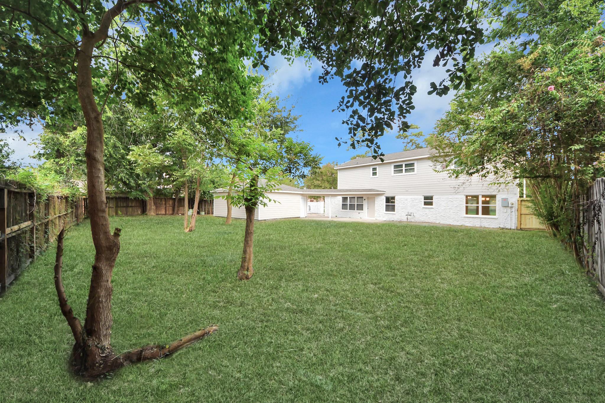 White two-story house and extension at the far end of a fenced, tree-shaded grassy backyard.