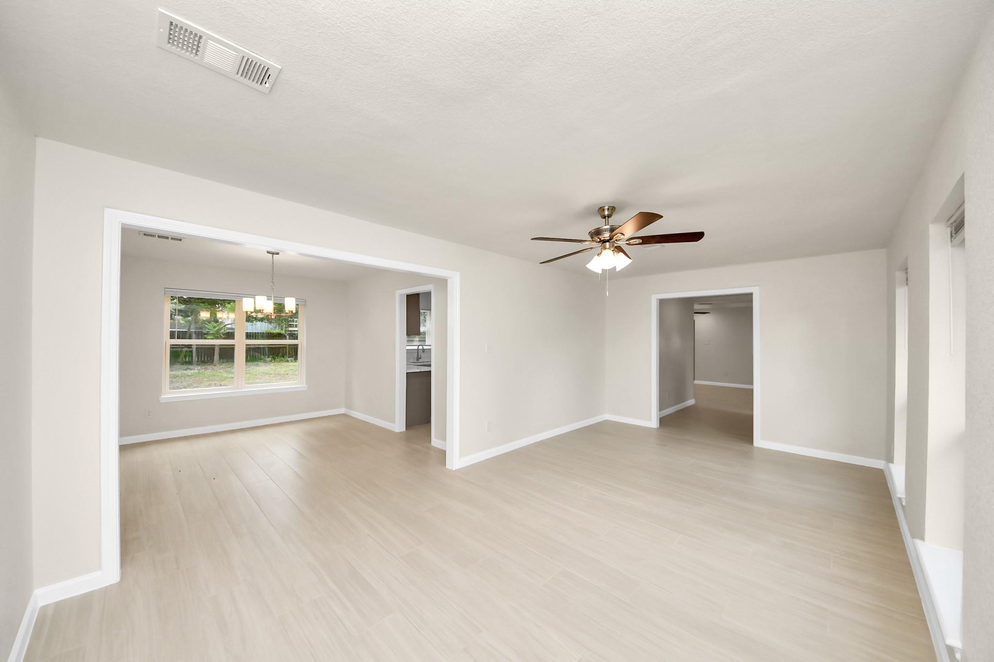 Empty living room with pale tile floors, ceiling fan, and a wide opening to a windowed dining area.