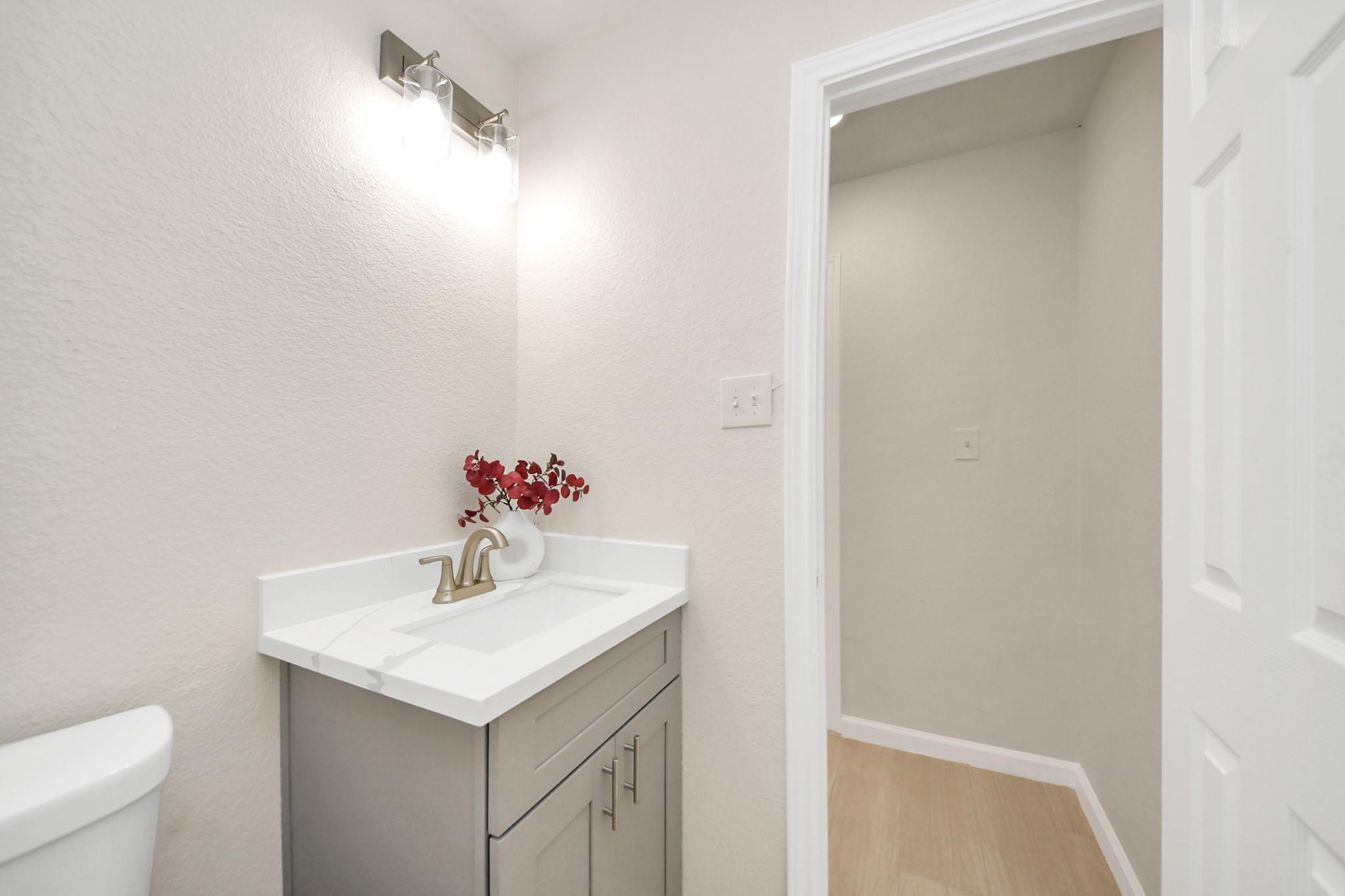 Bathroom vanity with a white countertop, gray cabinet, gold-tone faucet, and red flowers beside an open doorway.