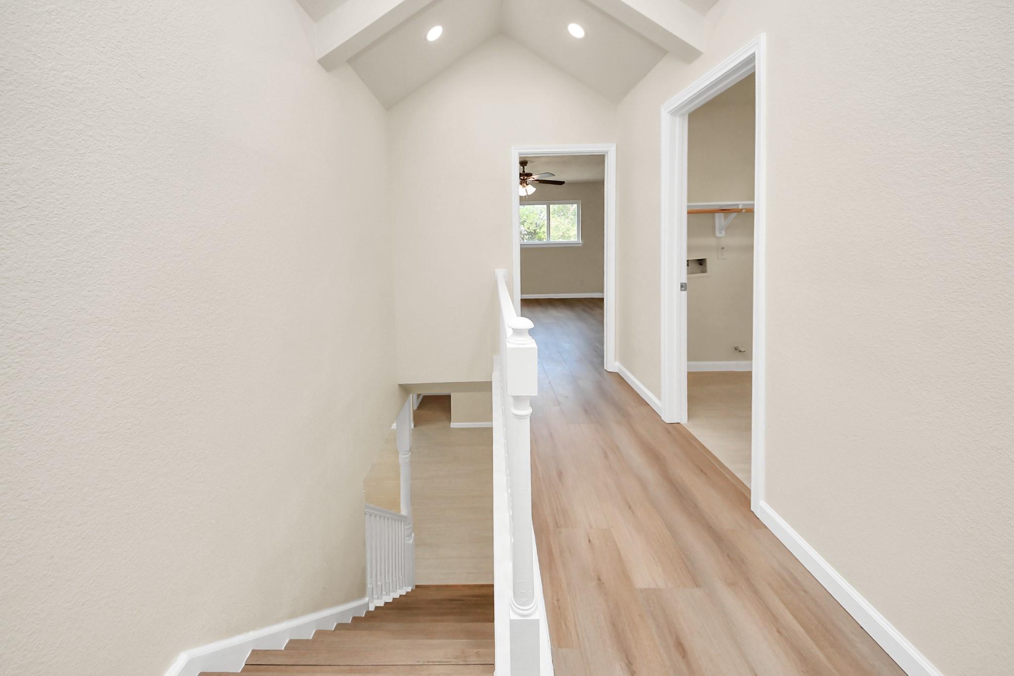 Empty upstairs hallway with vaulted ceiling, light wood floors, and stairs descending beside the landing.