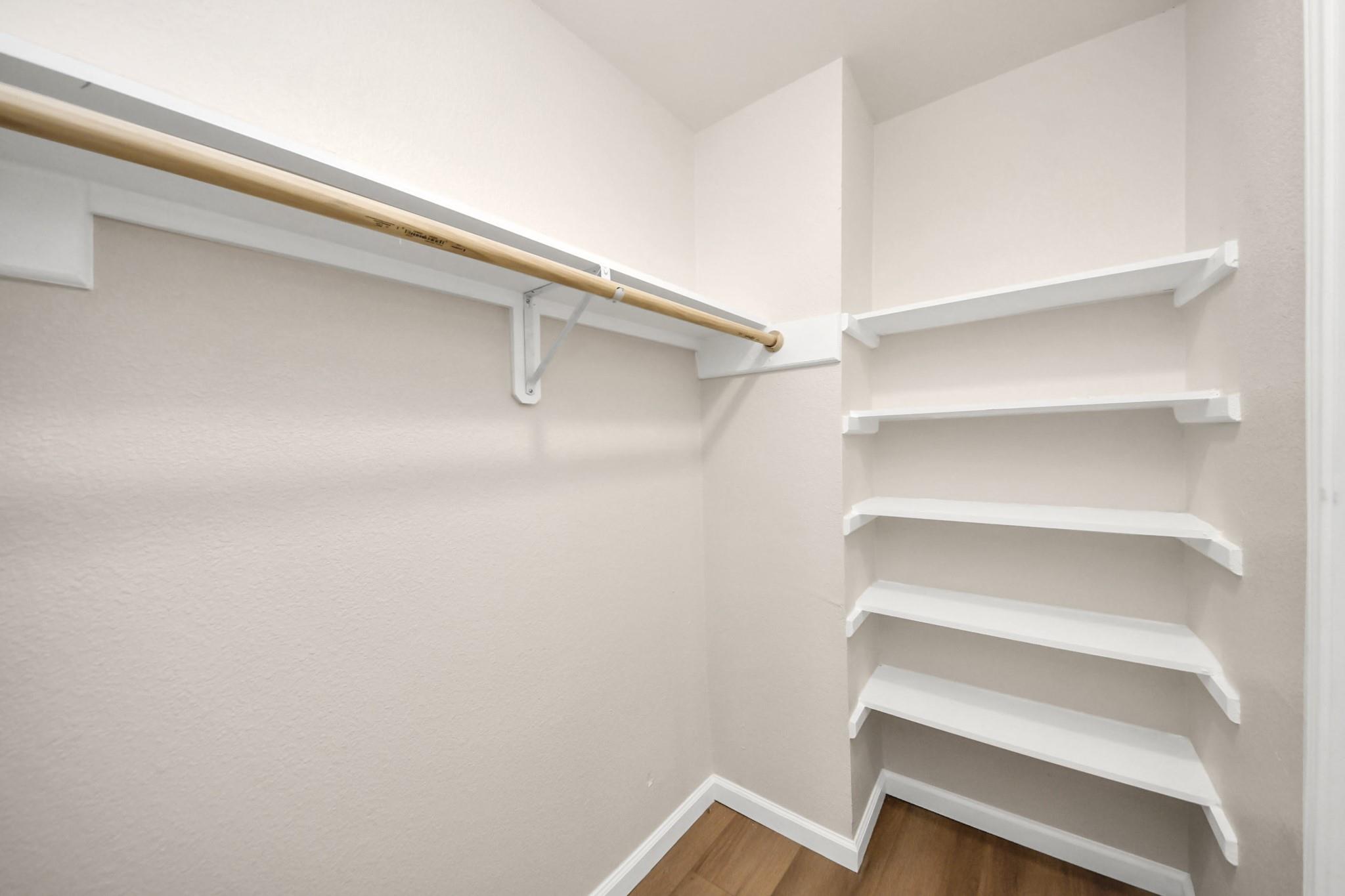 Empty walk-in closet with white built-in shelves, a wooden hanging rod, and wood flooring.