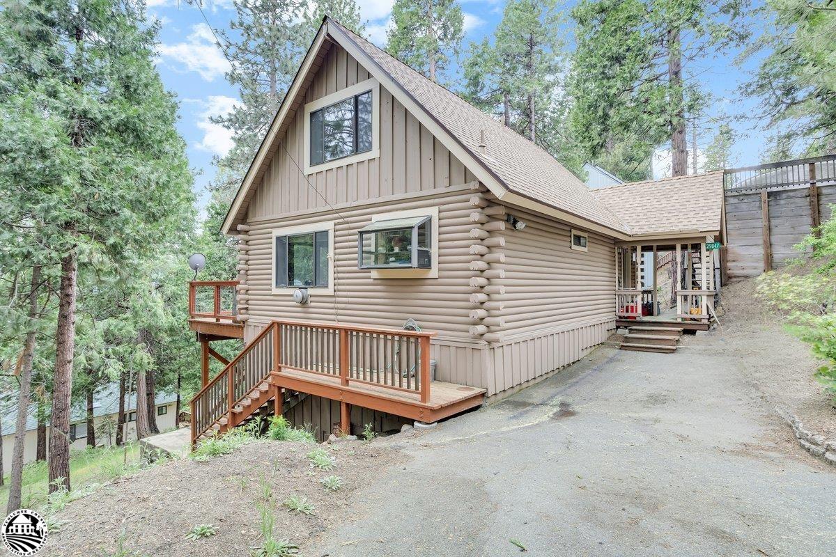 Log-sided cabin among pine trees, with a raised wooden deck and stairs beside the driveway.