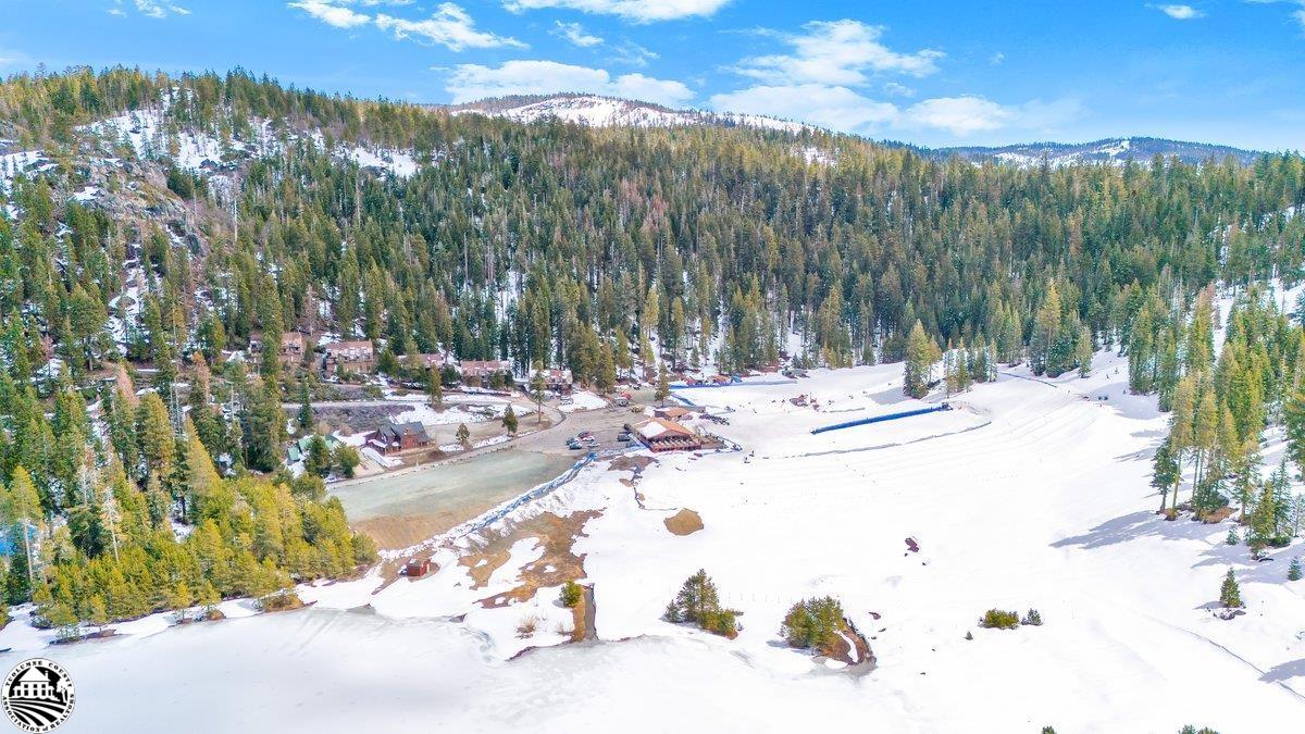 Snow-covered mountain meadow and pond surrounded by evergreen forest, with buildings and snowy ridges beyond.