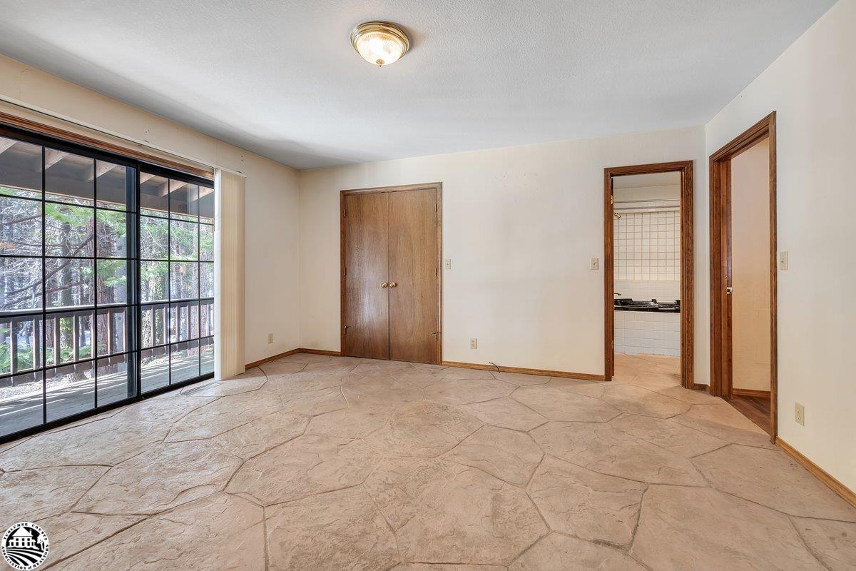 Empty tiled room with a large sliding glass door overlooking trees and a kitchen doorway.