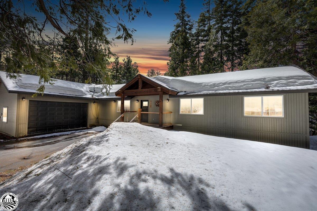 Snow-covered gray home with a double garage and timber entry porch among trees at dusk.