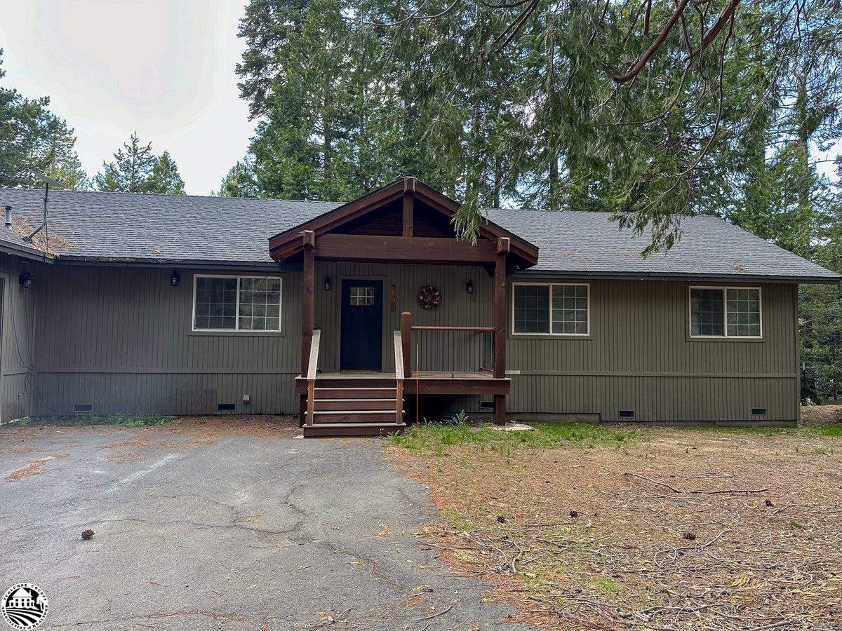 Gray single-story home with a dark wood covered porch, evergreen trees, and a paved driveway.