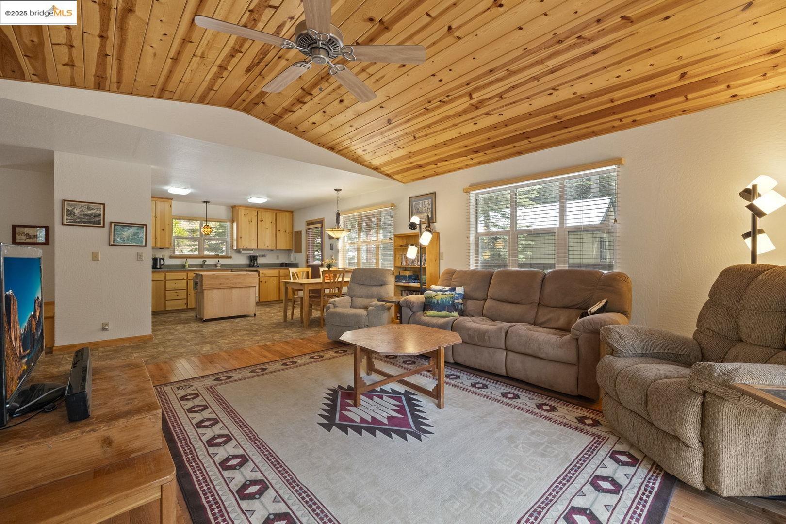 Living room with vaulted wood ceiling, large windows, and open kitchen and dining area.