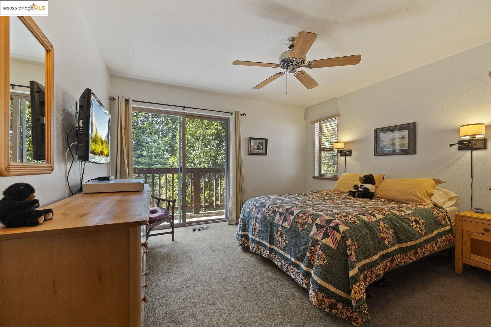 Bedroom with a patterned quilt and ceiling fan, opening onto a wooded deck through sliding glass doors.
