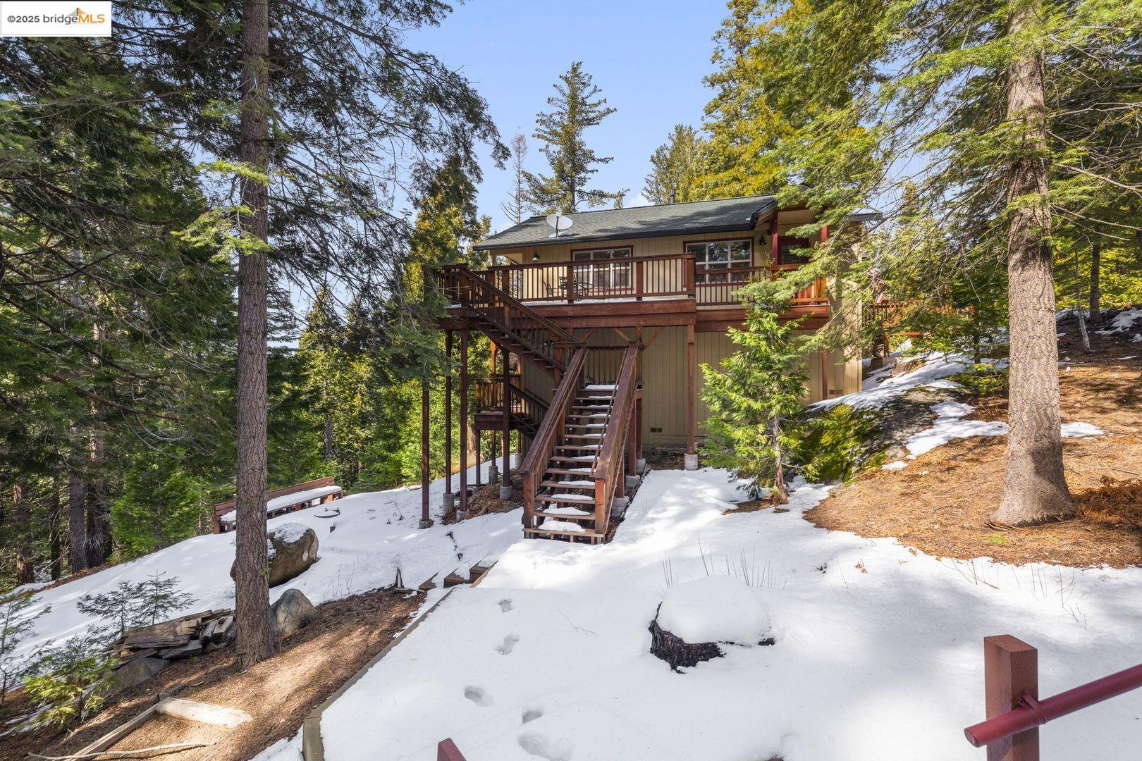 Wooded mountain home with an elevated wooden deck and stairs above a snow-covered yard.