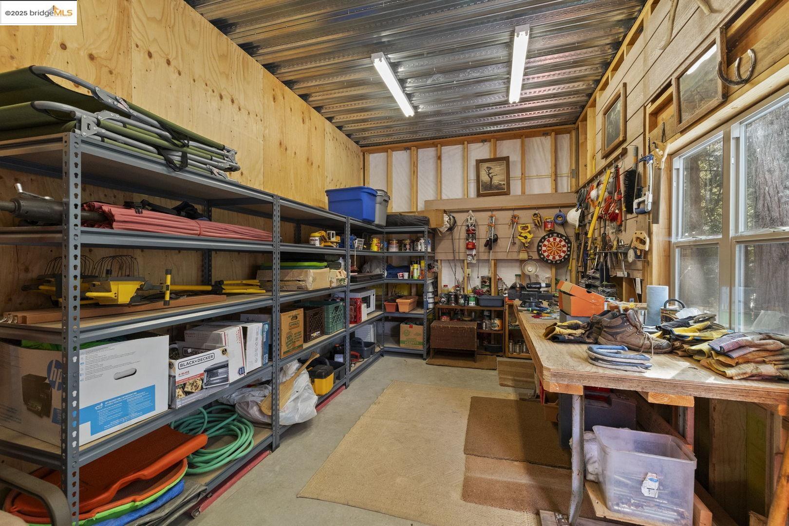 Workshop with storage shelves and a tool-covered workbench beside large windows.