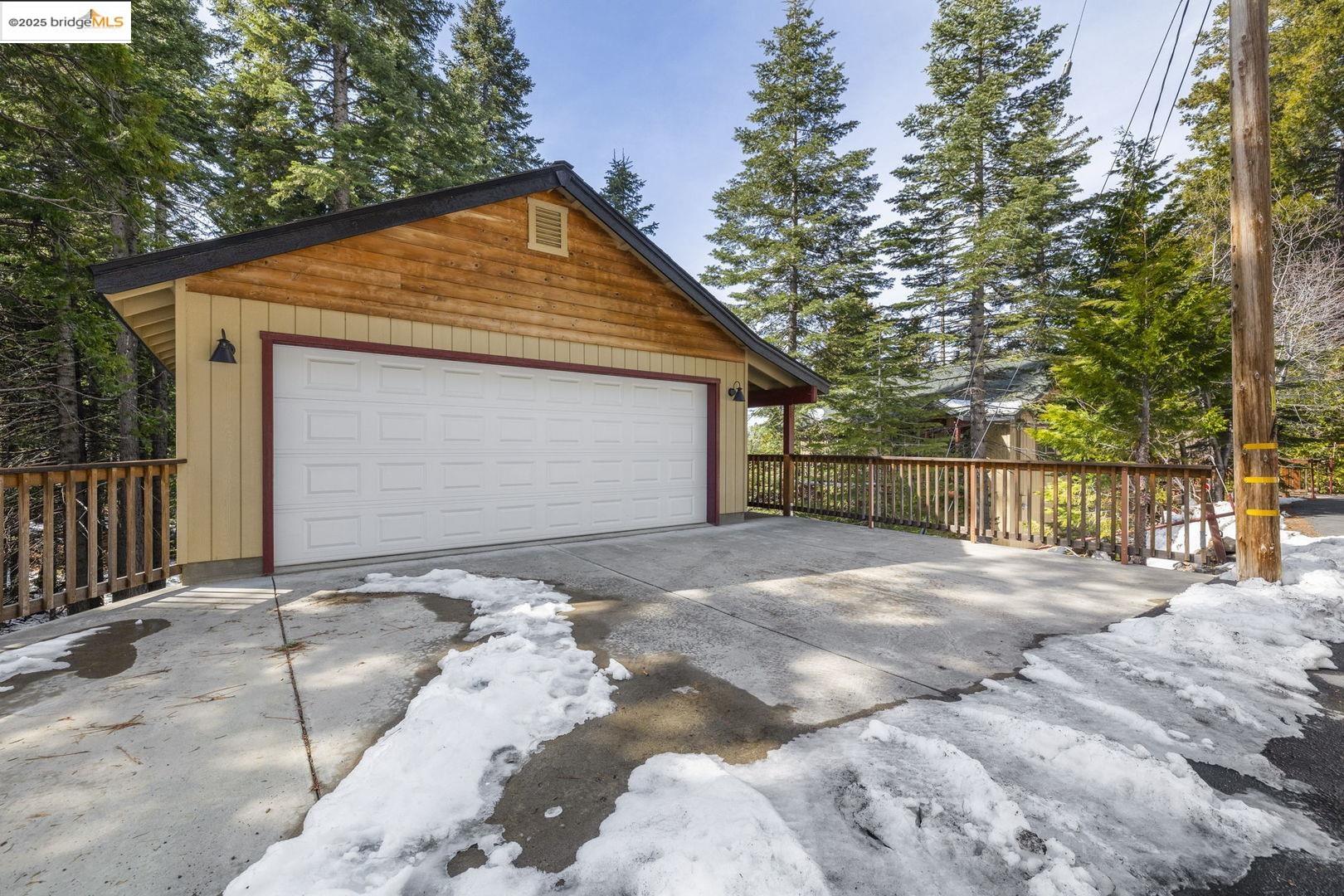 Detached wooden garage with a white double door, concrete driveway, and snow among evergreen trees.
